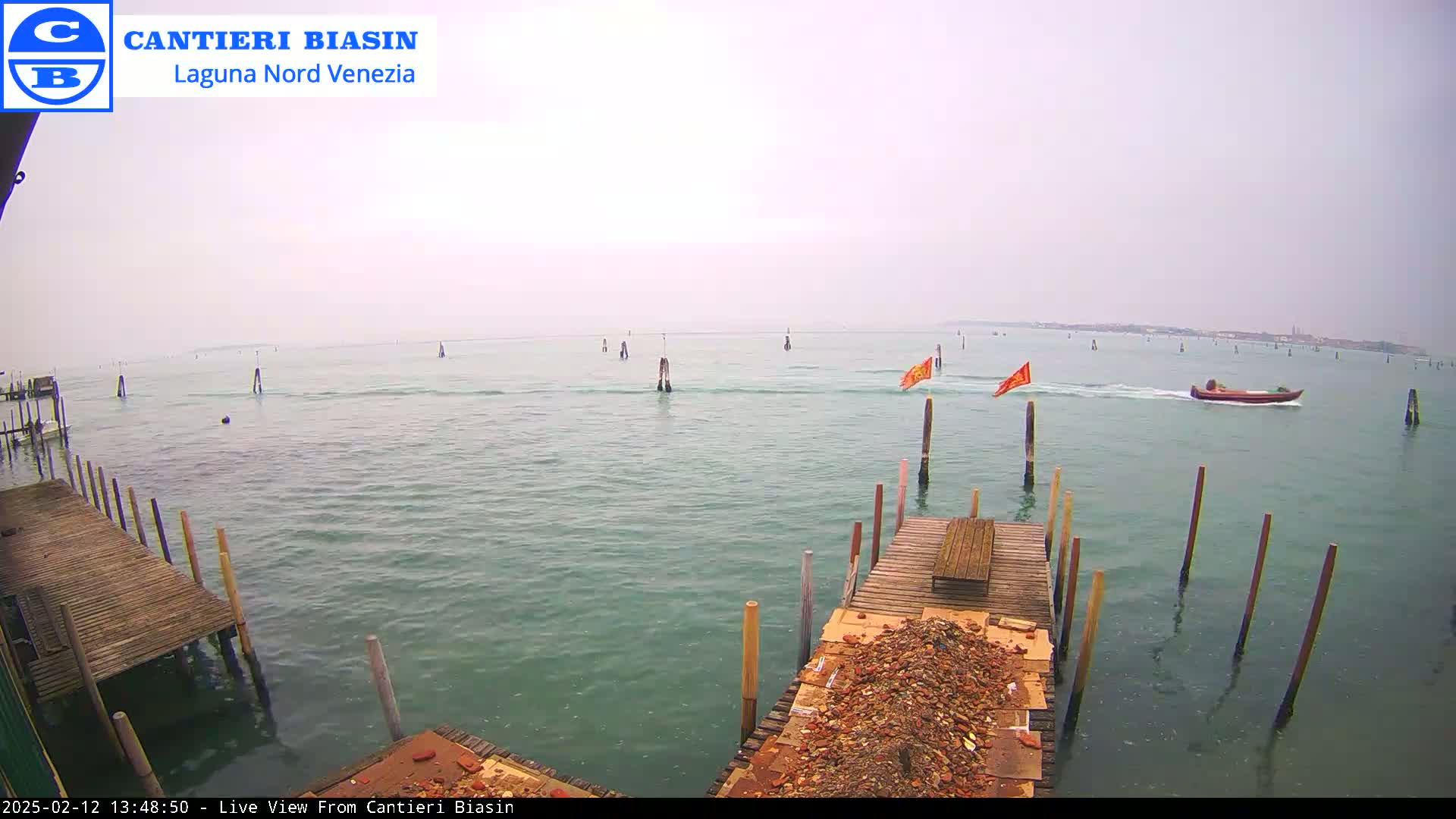A mostly cloudy day shows a body of water with a small motorboat, several wooden posts in the water, and a wooden dock with debris on it.