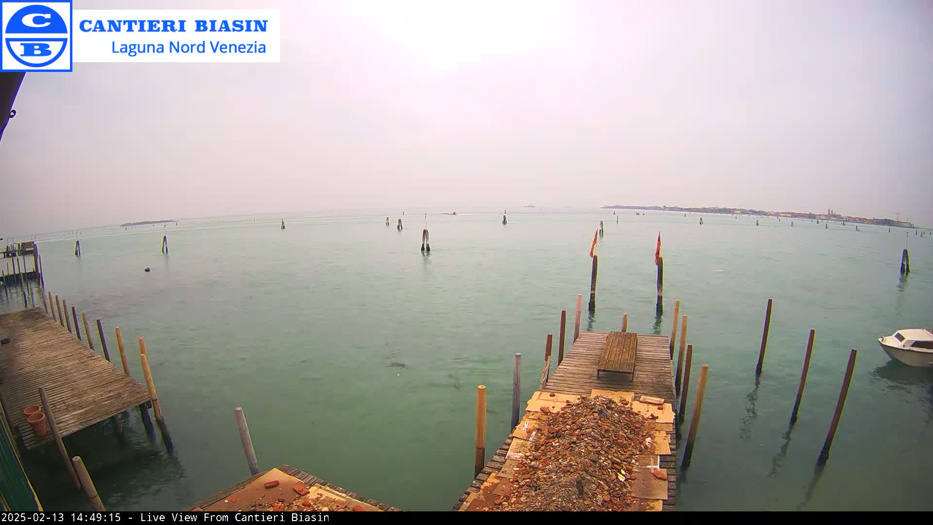 A hazy view of calm, green water shows wooden docks with piles of bricks and a small boat in the distance, under an overcast sky.