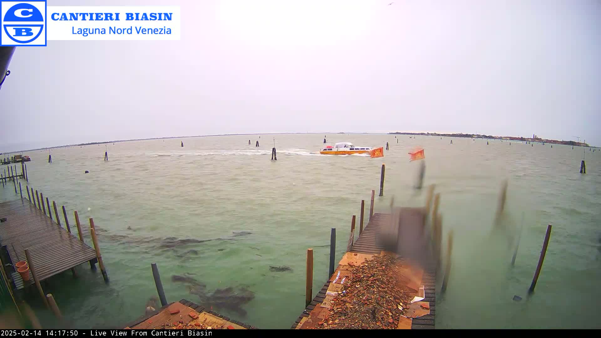 A yellow watercraft travels across a choppy body of water under an overcast sky, with wooden docks and pilings visible in the foreground.
