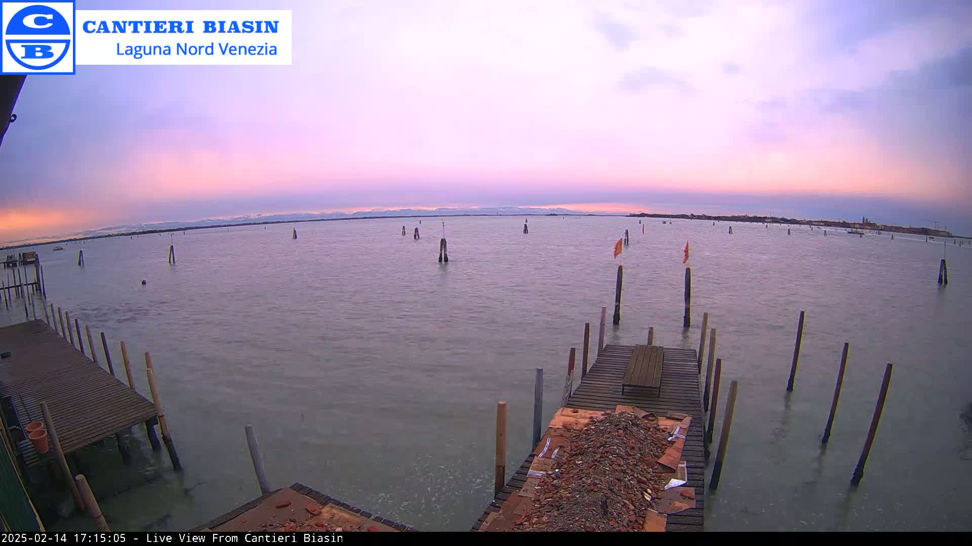 A calm body of water with numerous wooden posts extends to a distant shoreline under a pale pink and purple sunset sky.