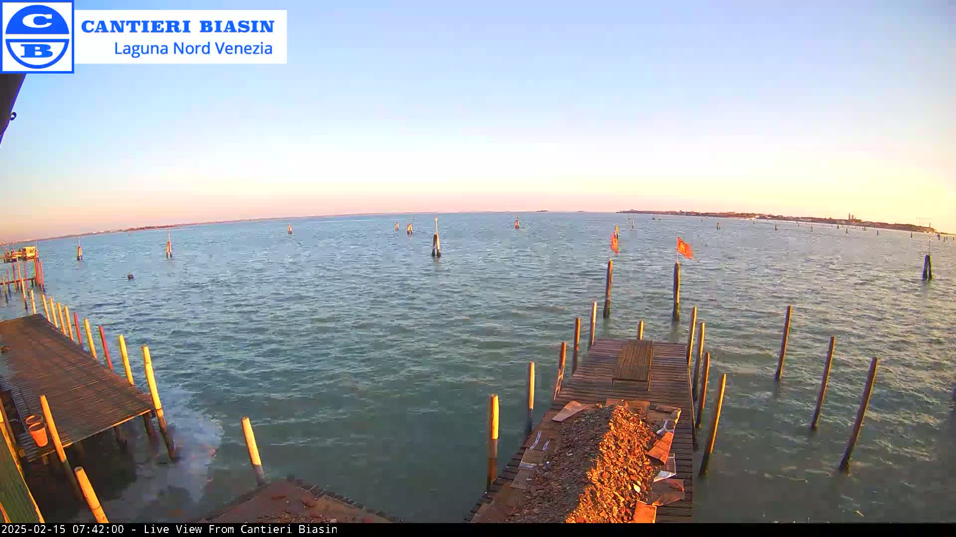 A calm body of water at sunset is seen from a wooden dock with several posts extending into the water.