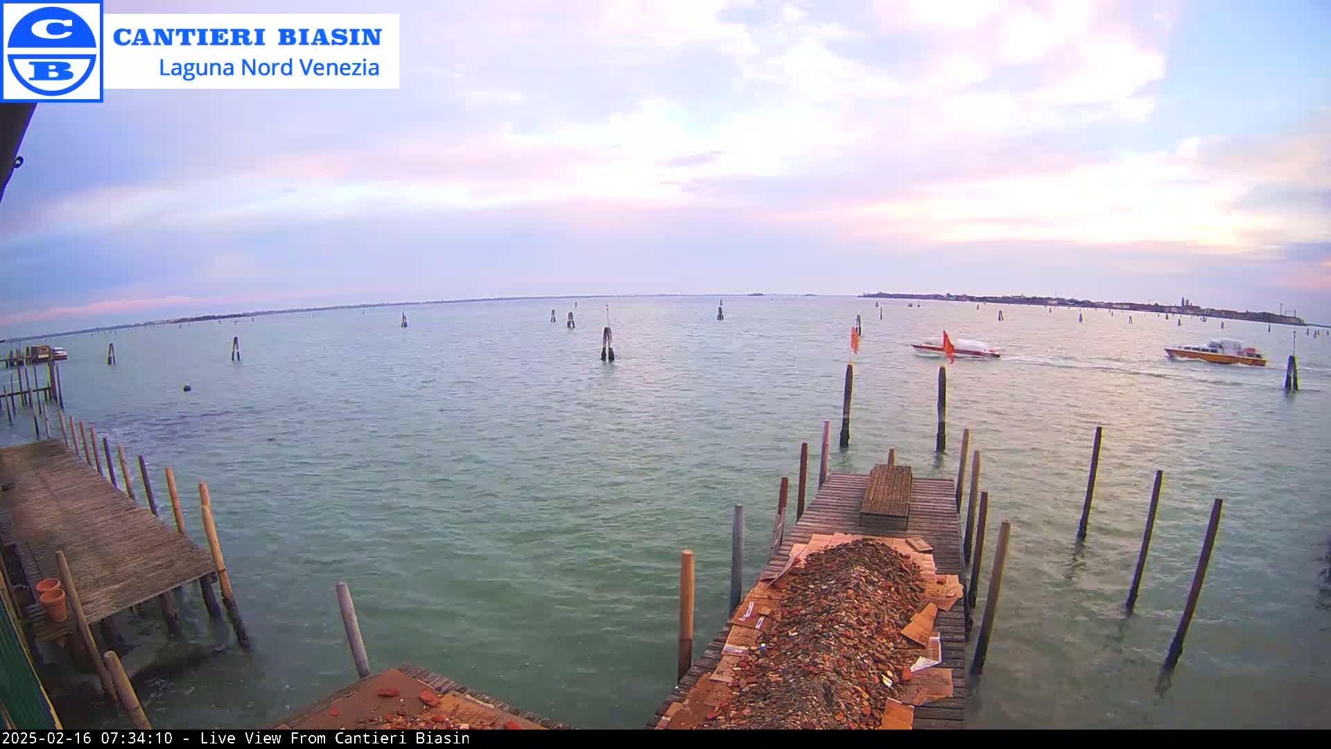 A calm body of water under a partly cloudy, pastel sky is viewed from a wooden dock with a pile of debris, featuring several boats and wooden posts in the water.