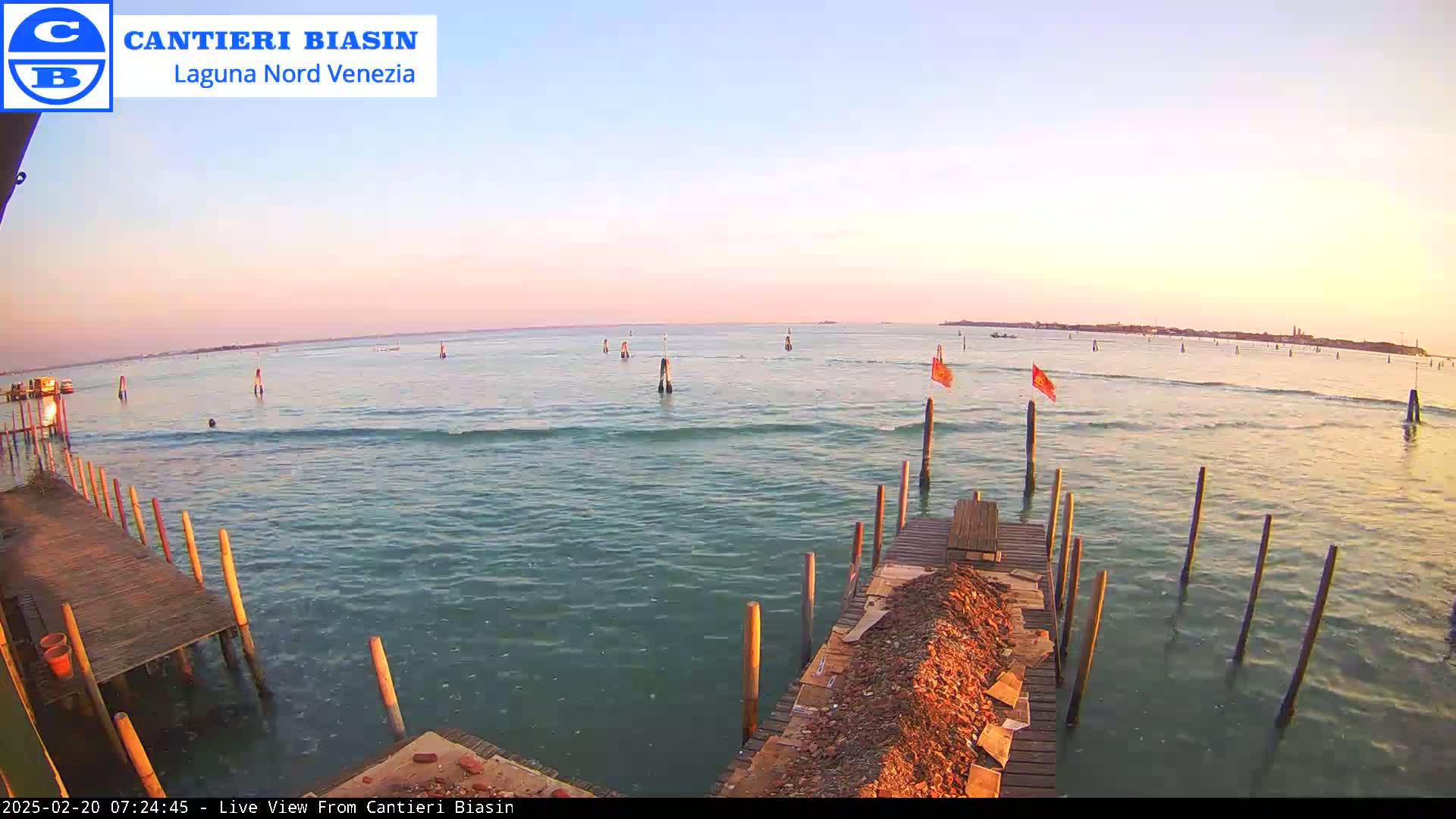 A calm body of water at sunset, showing a wooden dock and several posts extending into the water, with a distant landmass visible on the horizon.