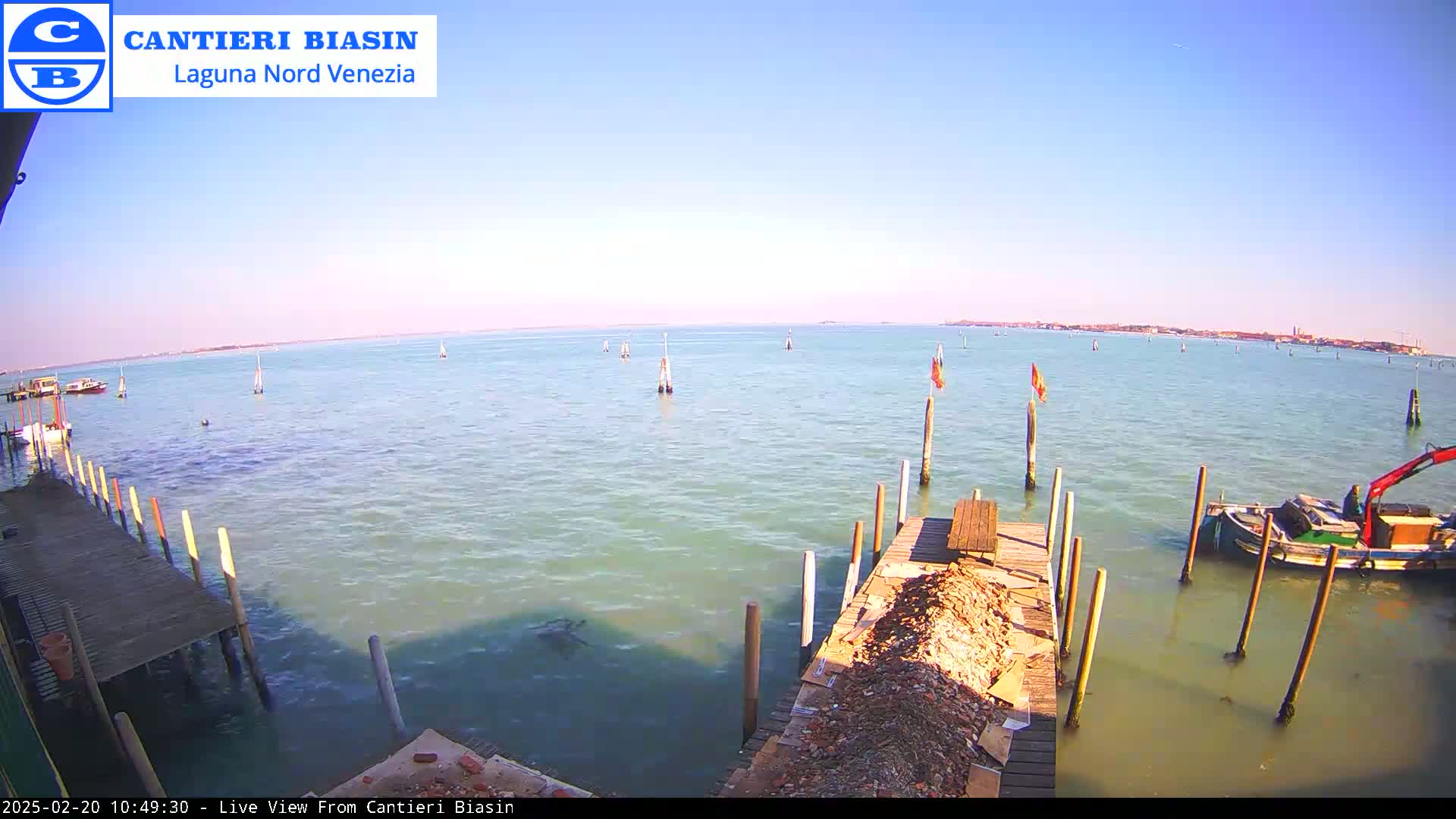 A calm, sunny day shows a body of water with several boats, a distant shoreline, and a wooden dock with a pile of debris.