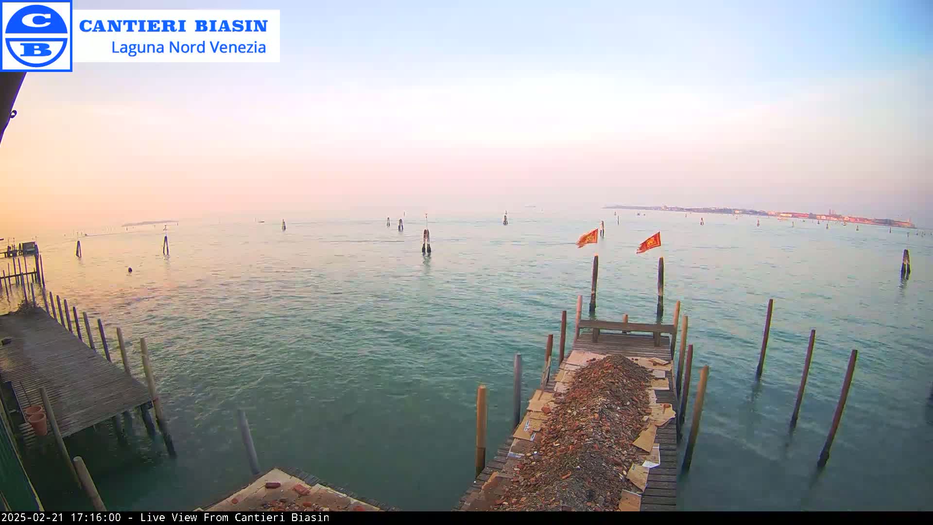 A calm body of water under a pale pink sky shows a wooden dock with a pile of debris, several posts in the water, and a distant landmass.