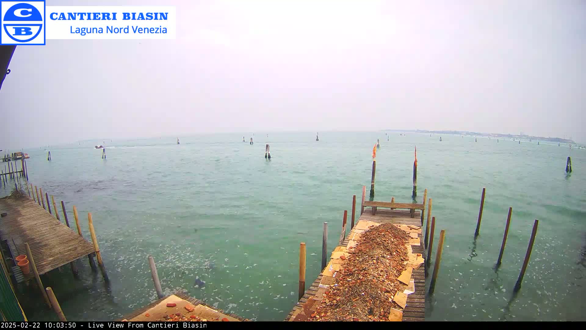 A calm, overcast day shows a body of water with several wooden posts and a wooden dock piled with debris in the foreground.
