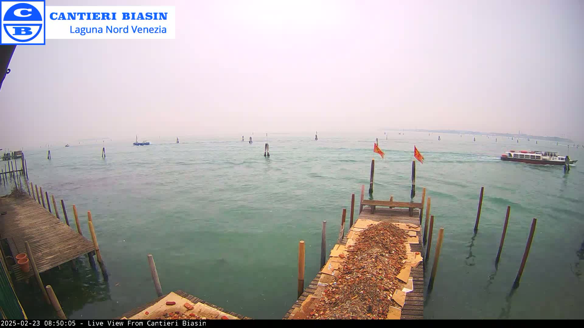 A hazy day shows a waterway with several boats, pilings, and a dock piled with debris, all viewed from a wooden dock.