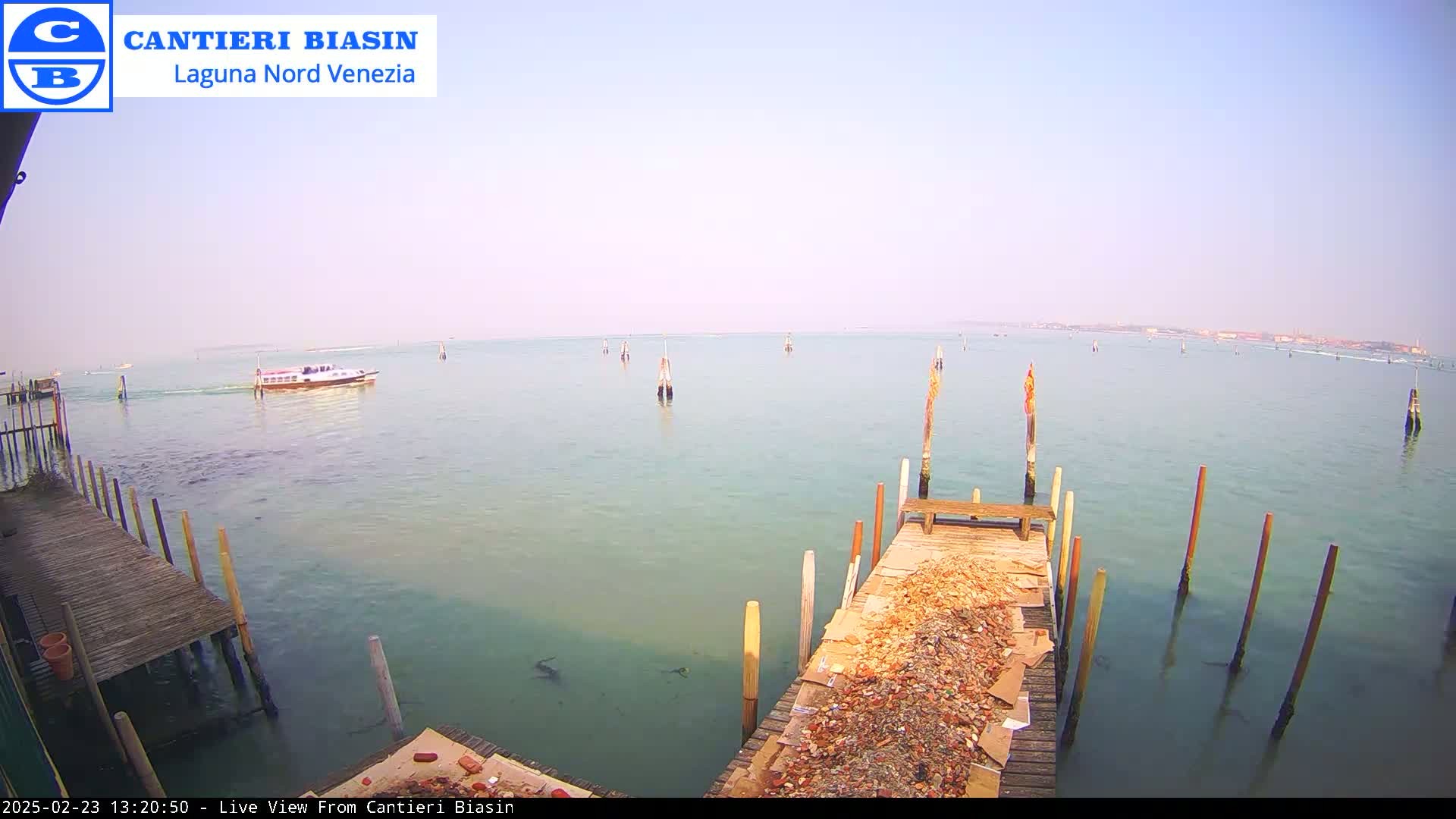 A calm, light-blue body of water is seen from a wooden dock, with a boat in the distance under a hazy sky.