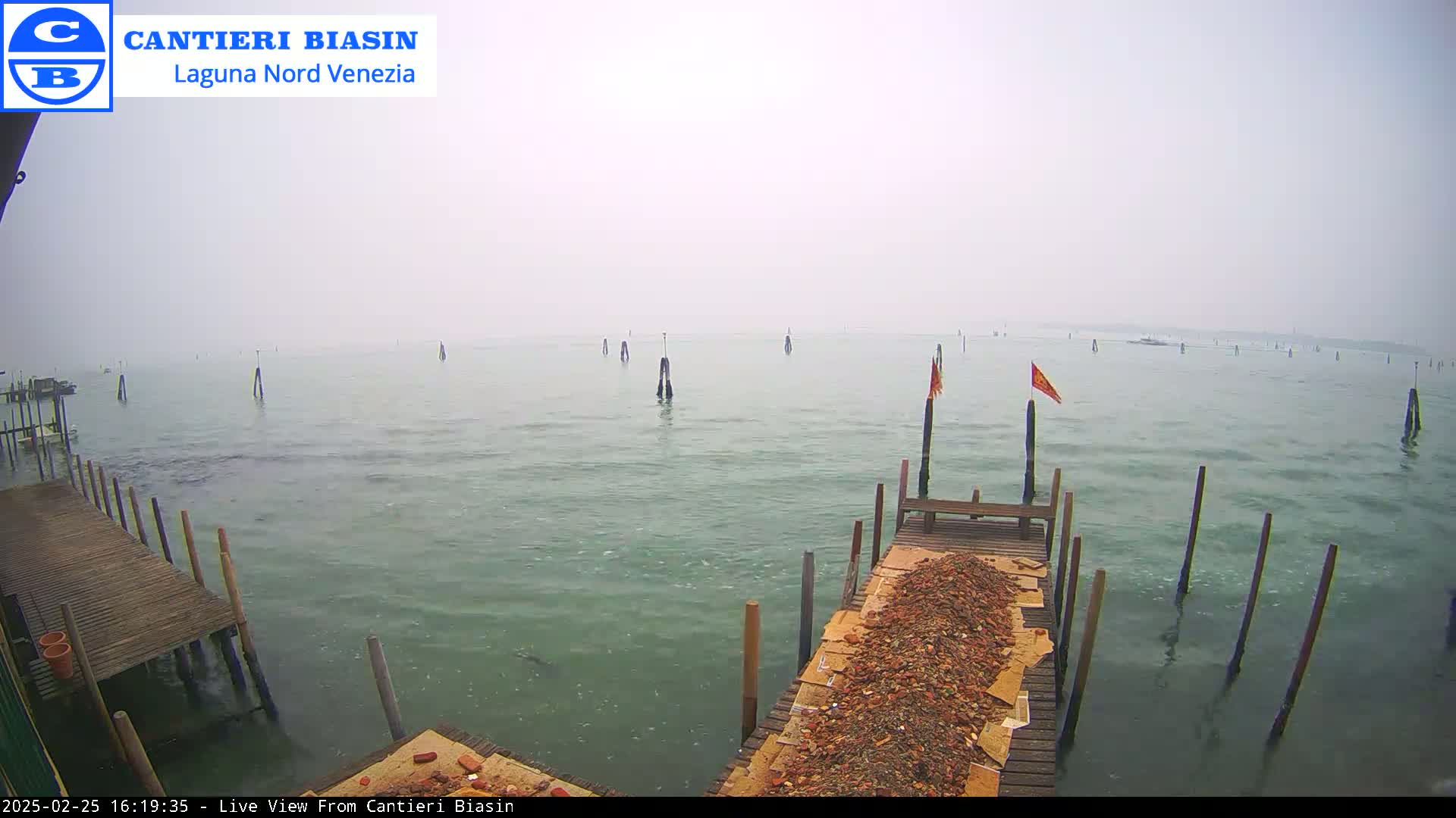 A hazy, overcast day reveals a calm body of water with several wooden posts and a small dock piled with debris, extending from a wooden structure.