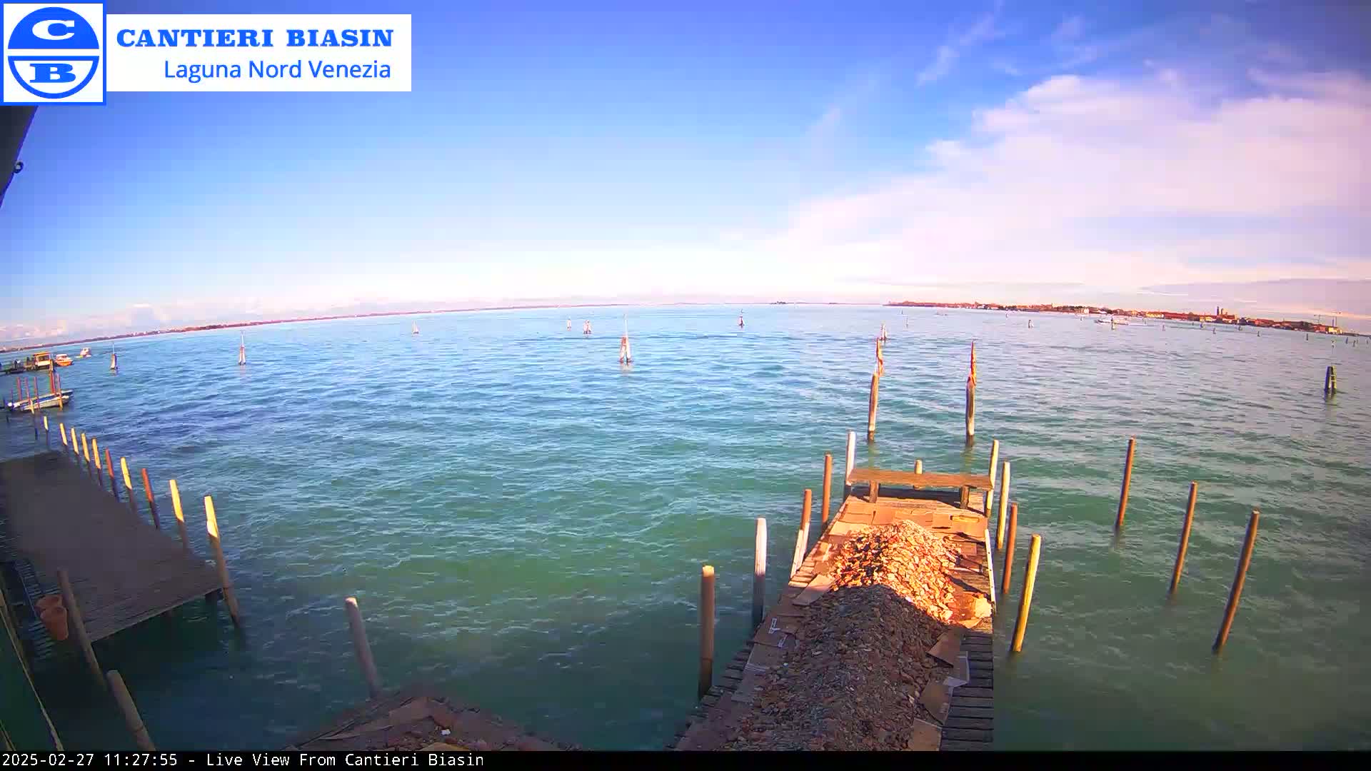 A mostly sunny day shows a calm body of water with several boats and pilings visible from a wooden dock.