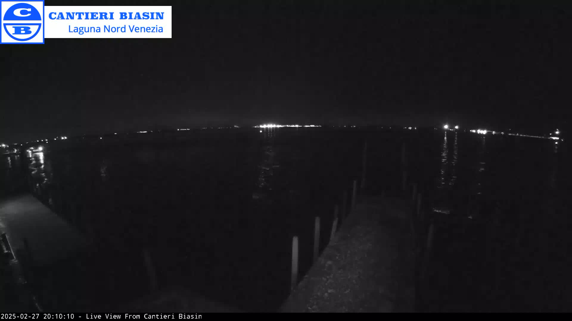 A nighttime view of a dark body of water with distant lights reflecting on the surface, seen from a pier or dock.