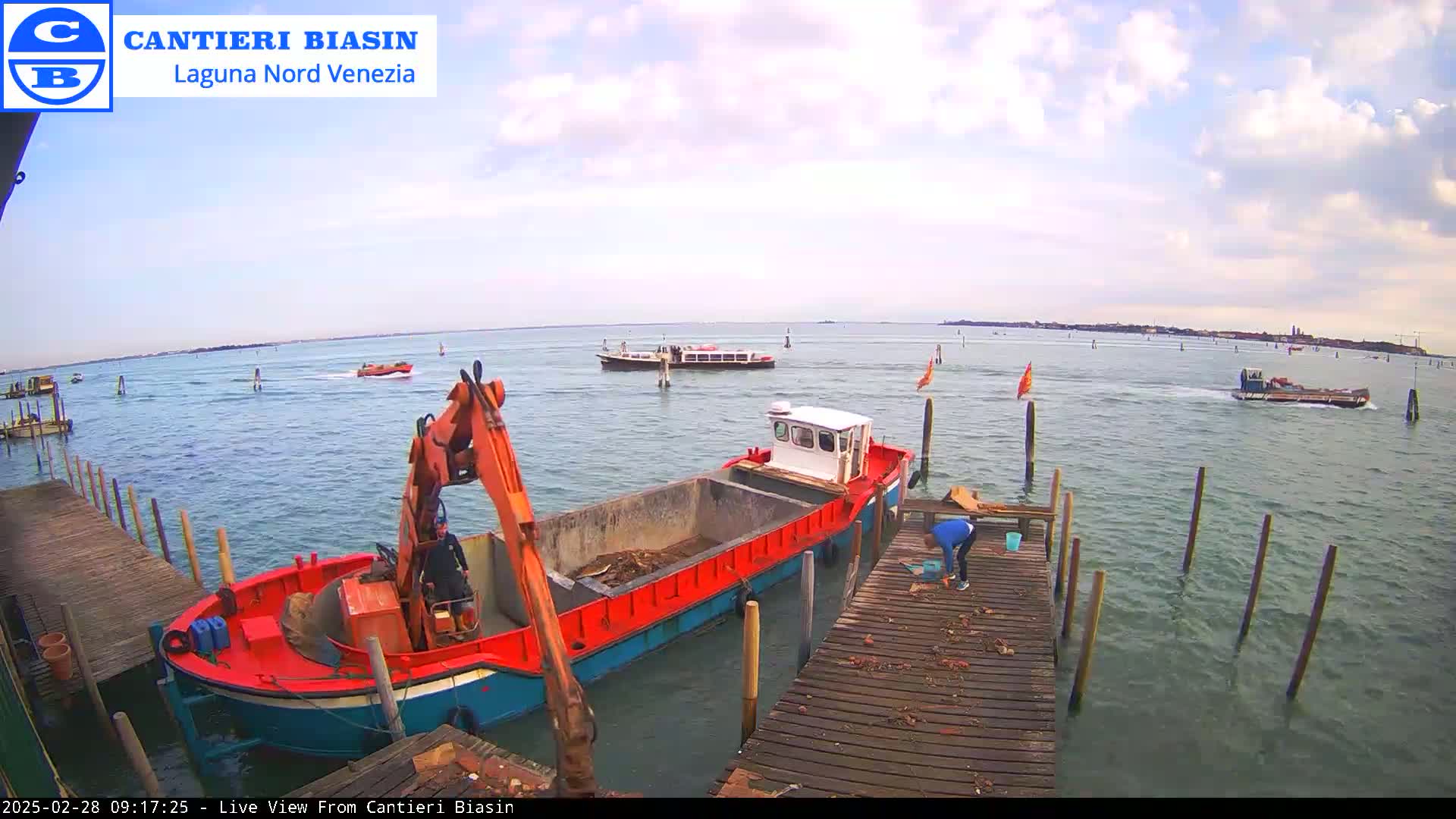 A red and blue barge loaded with debris is docked at a wooden pier, with a person working nearby, while other boats are visible on a calm body of water under a partly cloudy sky.