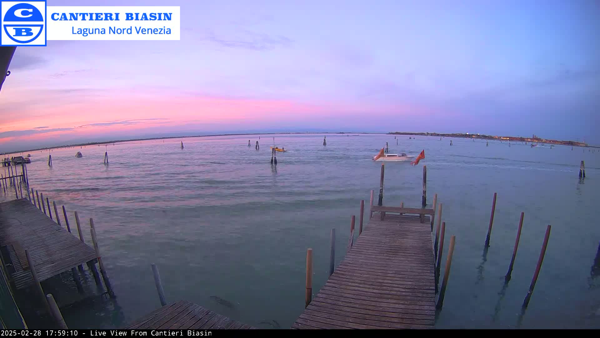 A calm body of water at sunset shows a wooden dock extending into the water with several boats and poles visible in the distance under a pale pink and purple sky.