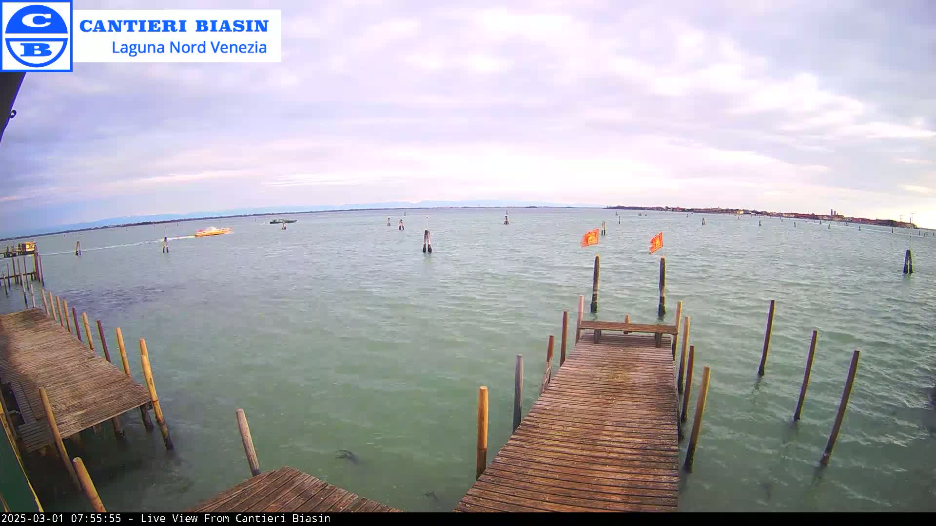 A wooden dock extends over calm, greenish-grey water under a cloudy sky, with a few boats visible in the distance and orange flags marking posts in the water.
