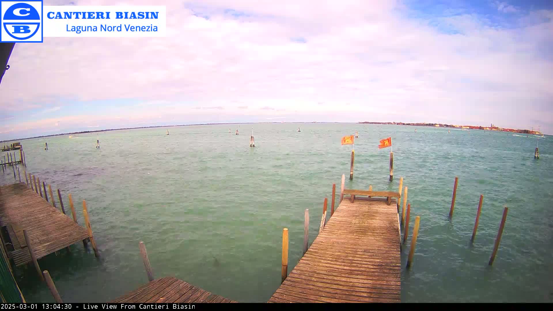 A wooden dock extends over greenish-blue water under a partly cloudy sky, with a distant shoreline visible.