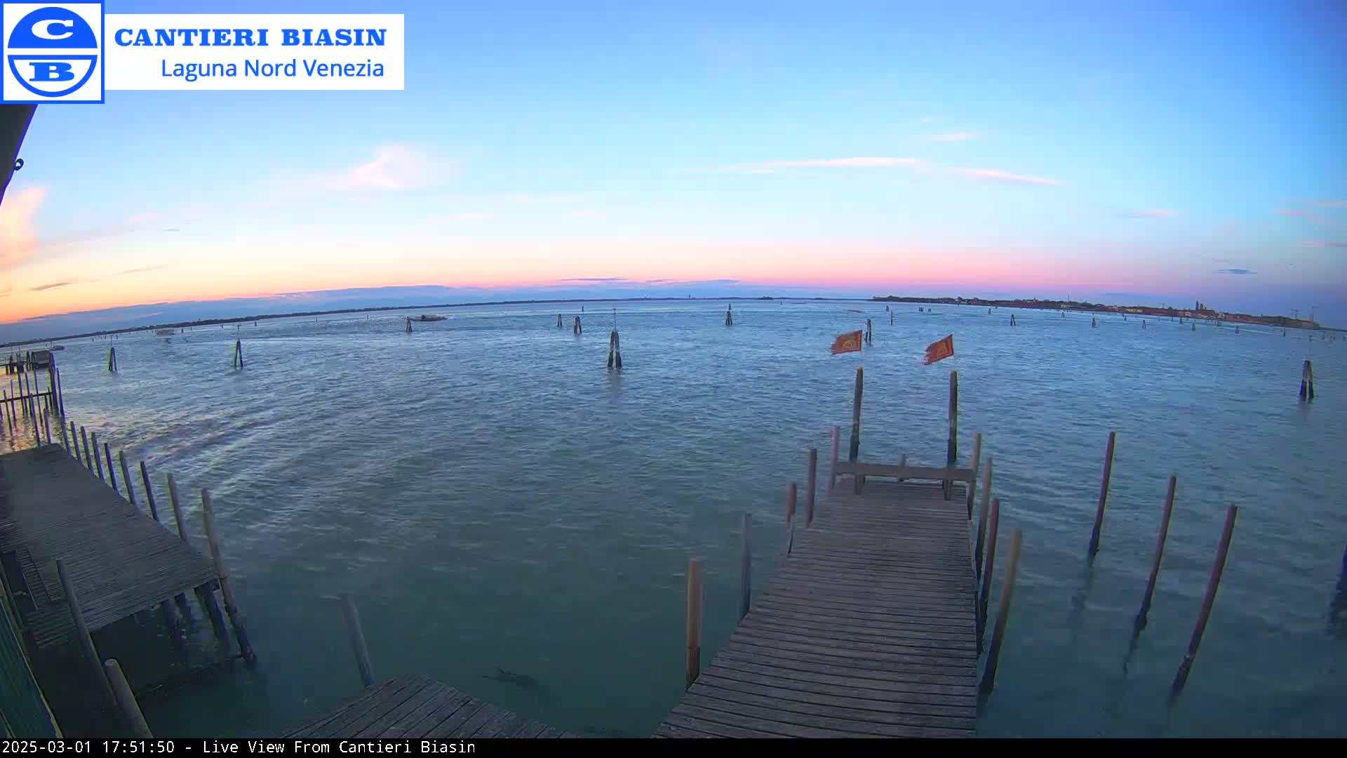 A wooden dock extends over calm water at sunset, with several posts and a few small flags visible in the distance.