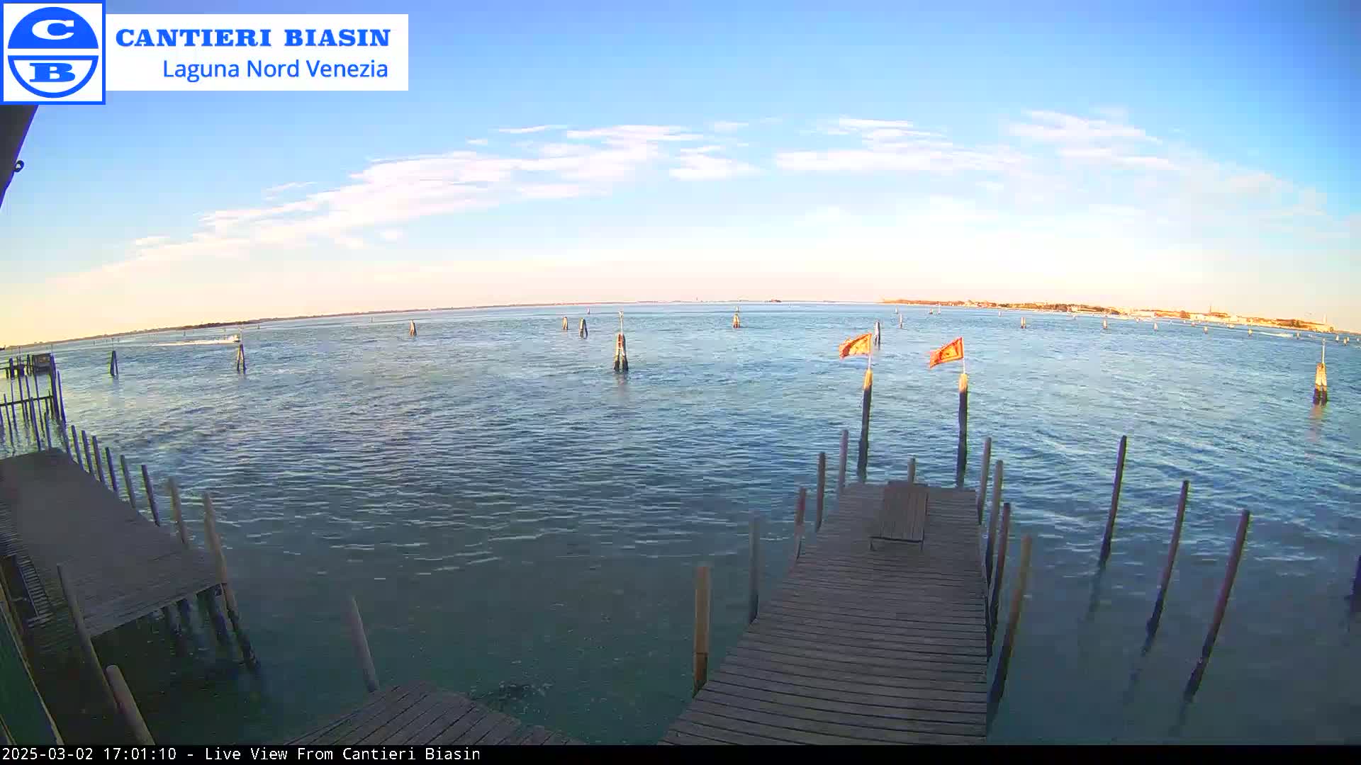 A wooden dock extends over calm, blue water under a mostly clear, light-blue sky; numerous posts and two orange flags are visible in the water.