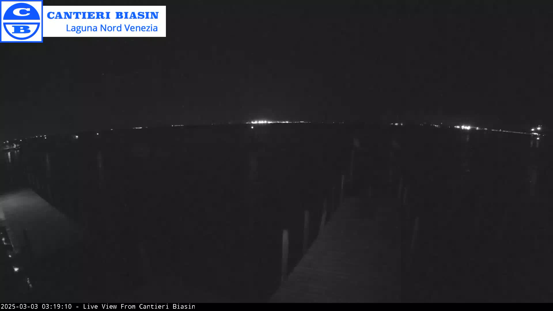 A nighttime view shows a dark body of water with distant lights on the horizon and a dark dock or pier in the foreground.
