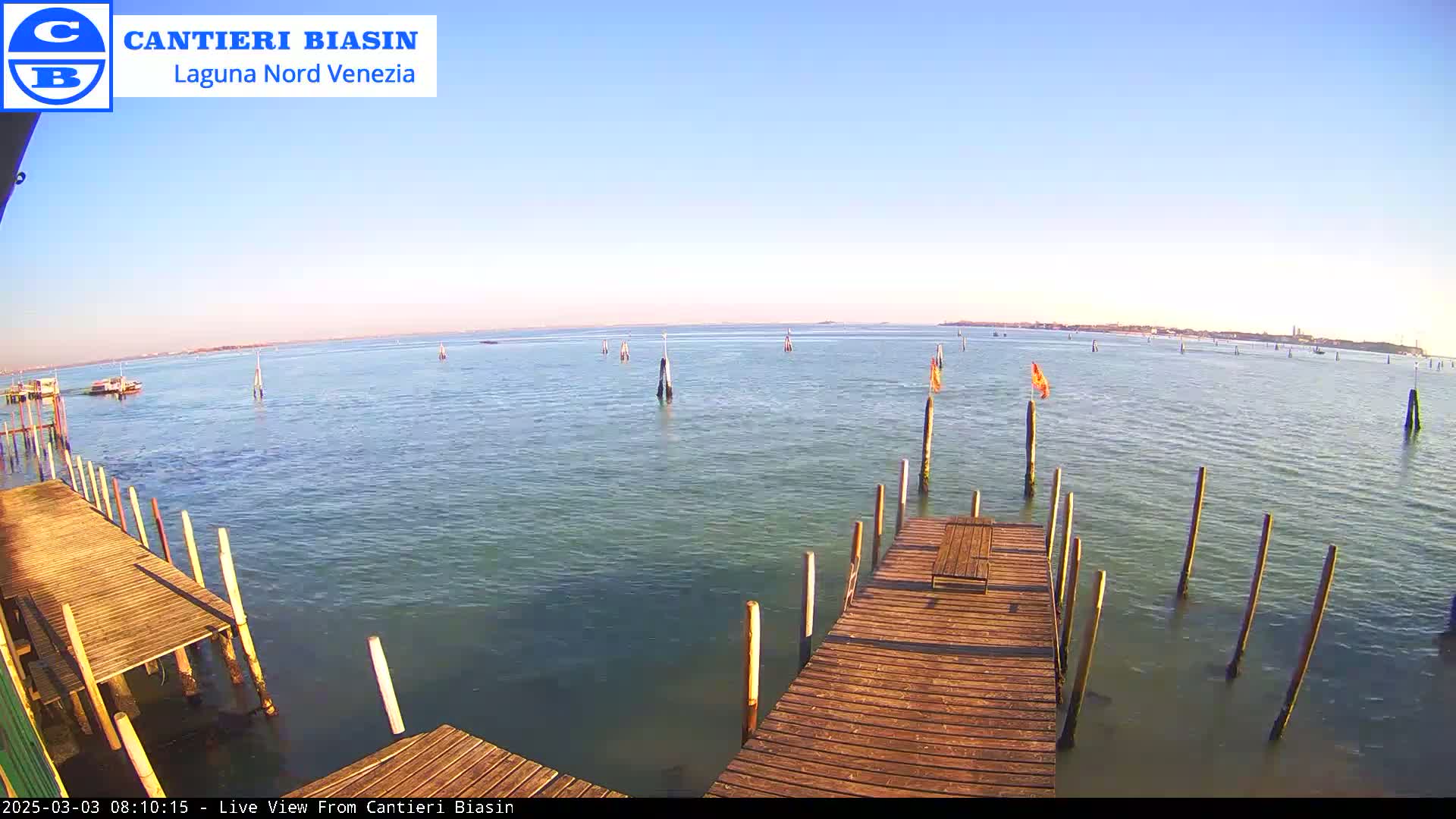 A wooden dock extends over calm, light-green water under a clear blue sky, with distant land visible on the horizon.