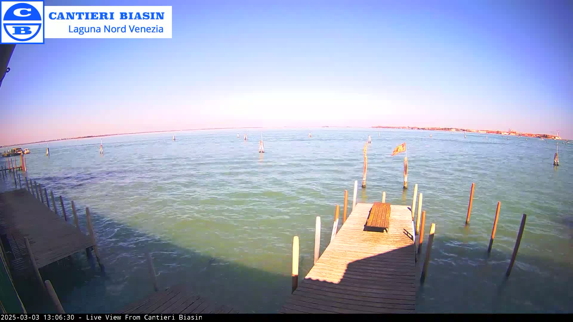 A wooden dock extends over calm, greenish-blue water under a clear, bright blue sky, with several posts in the water and a distant shoreline visible.