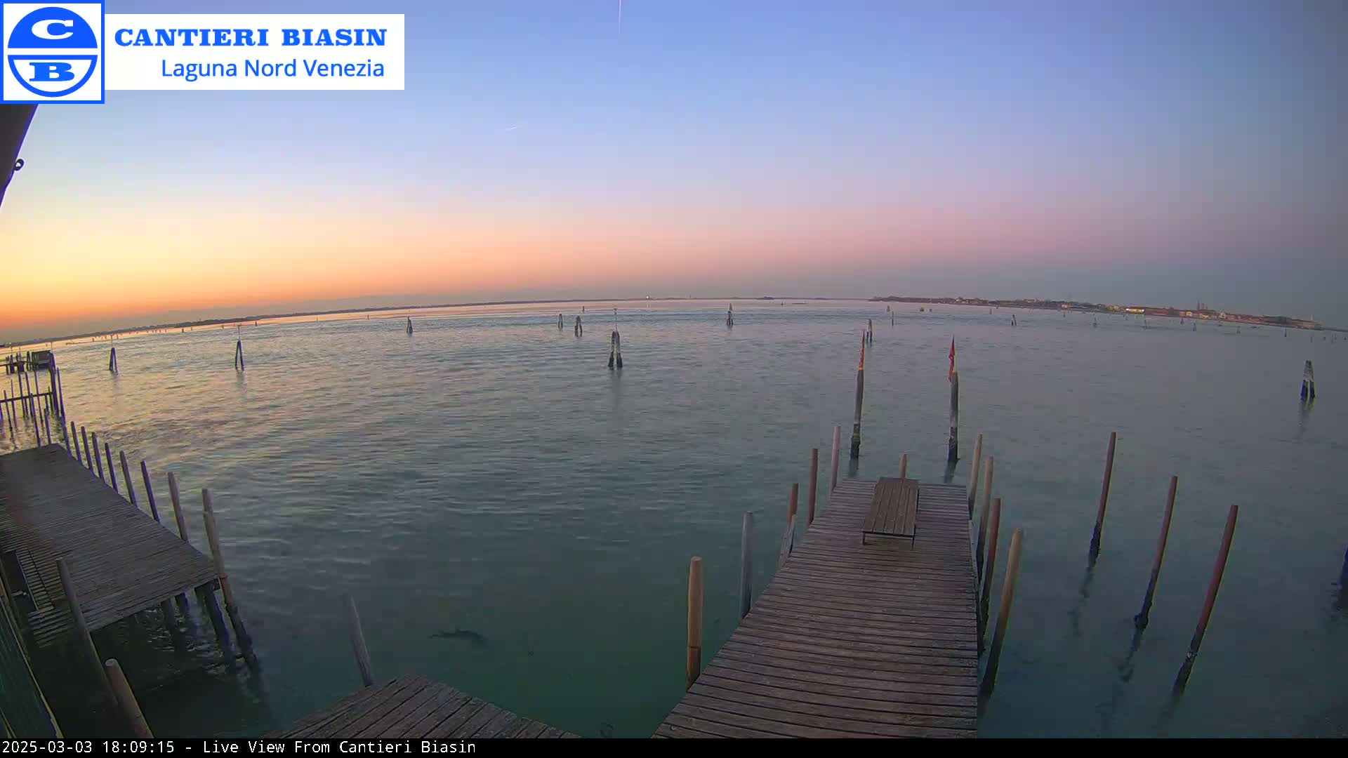 A tranquil body of water at sunset, showing a wooden dock extending into the calm water with several posts scattered throughout the scene.