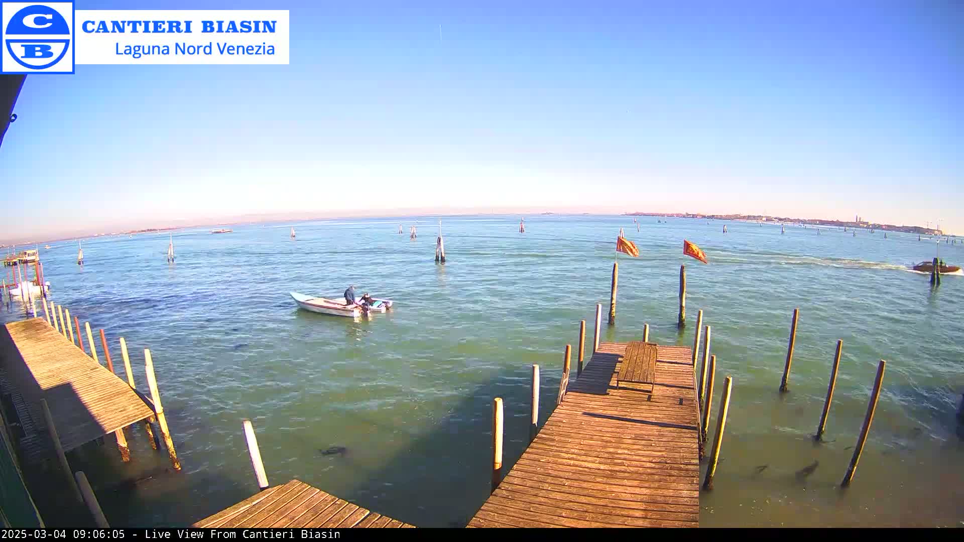 A sunny day shows a calm body of water with a small boat and several pilings extending from a wooden dock into the water, a distant shoreline visible in the background.