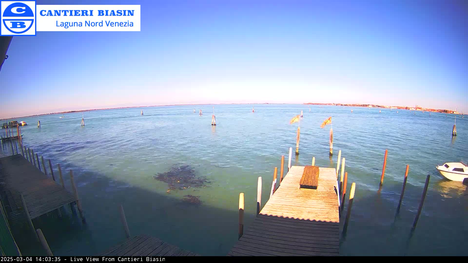 A wooden dock extends over calm, shallow, blue-green water under a clear blue sky, with a small boat and distant land visible in the background.