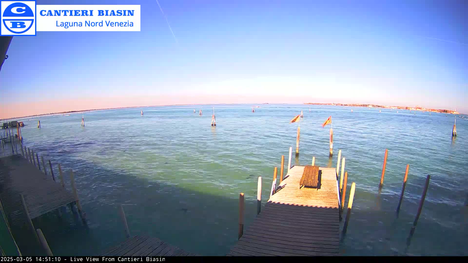 A wooden dock extends over calm, greenish water, with several posts and flags in the distance under a clear blue sky.