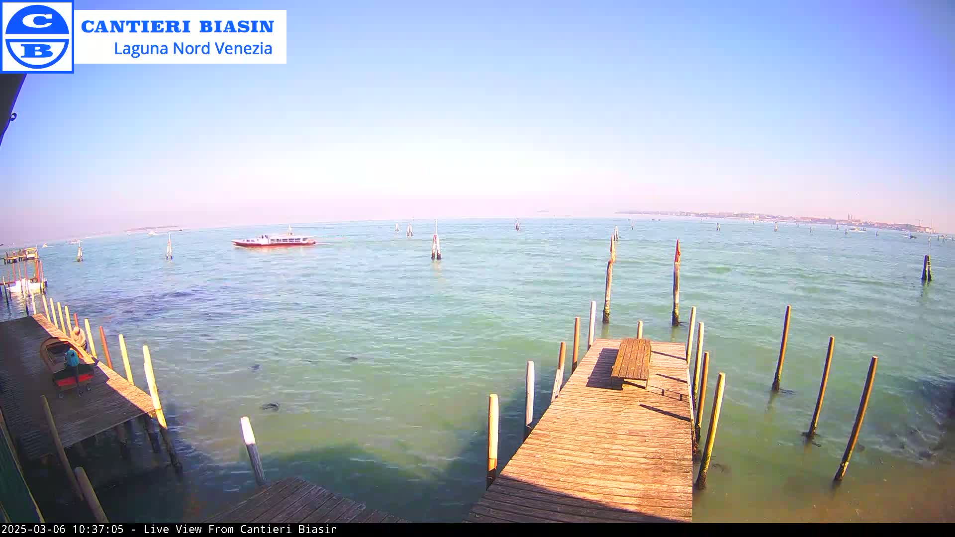 A wooden dock extends over calm, light-green water under a clear blue sky, with a boat visible in the distance and several wooden posts in the water.
