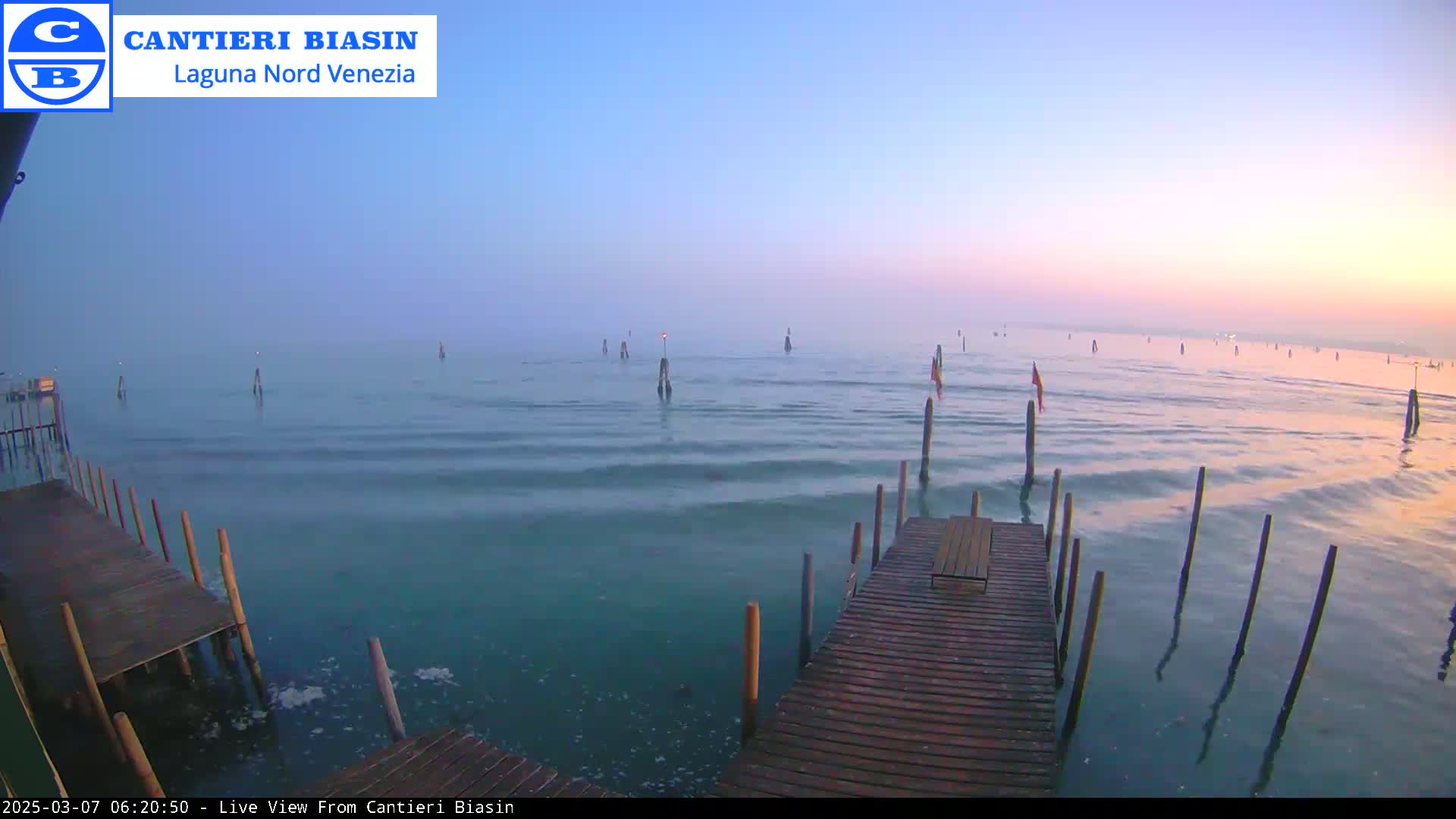 A wooden dock extends over calm, blue-green water under a pale pink and blue sunrise.