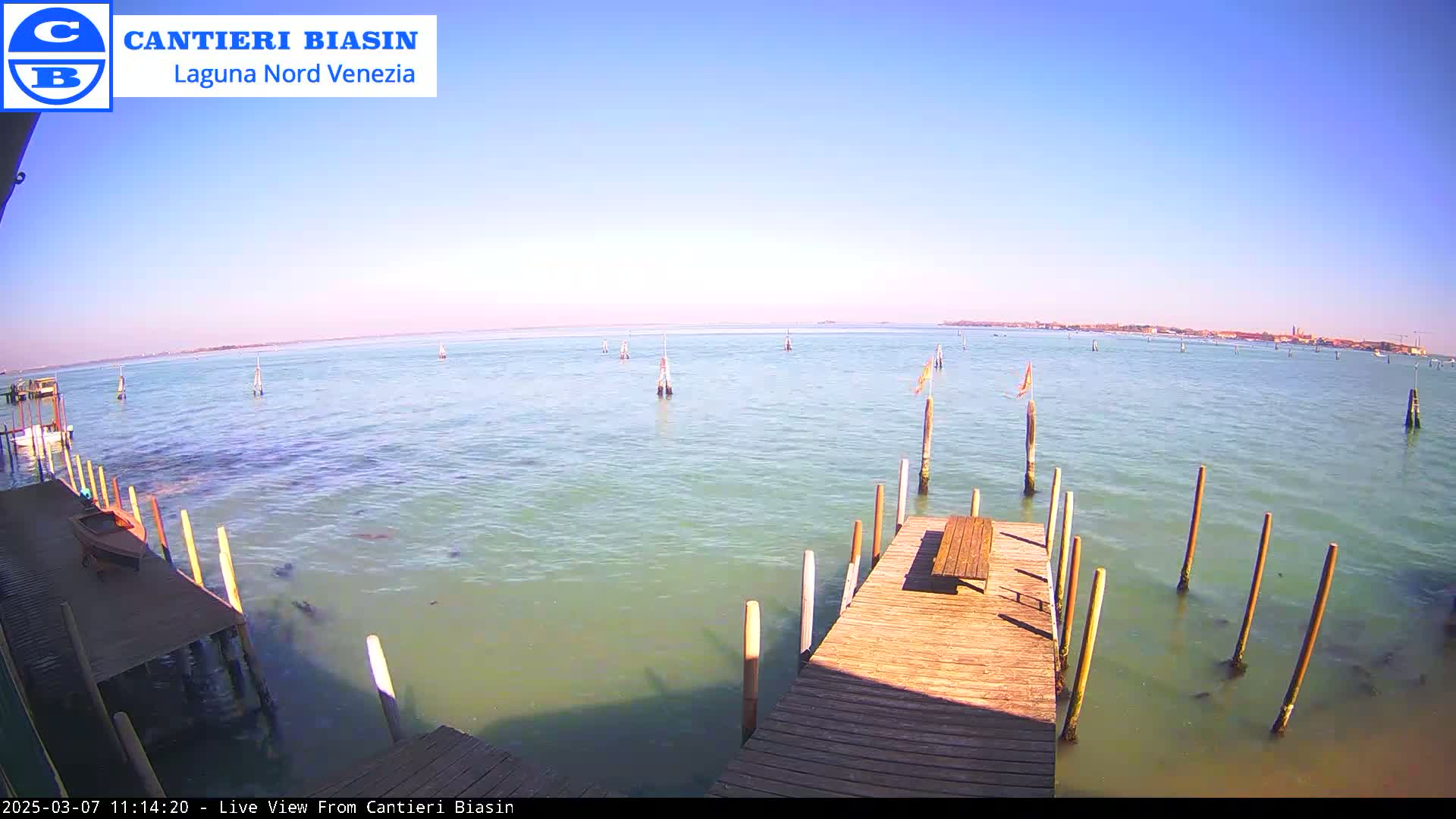 A wooden dock extends over calm, light-green water, toward a distant shoreline under a clear blue sky.