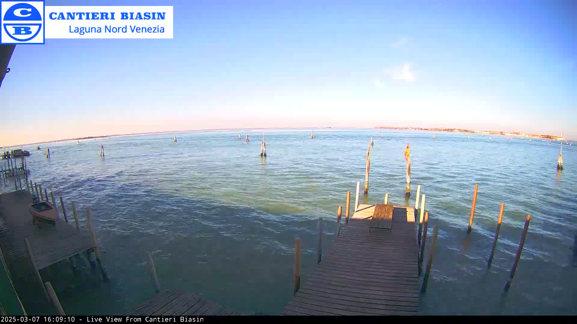 A wooden dock extends over calm, greenish-blue water under a clear blue sky, with numerous wooden posts in the water and a small boat moored nearby.