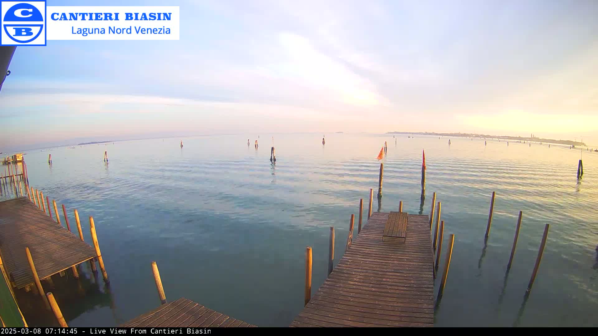 A wooden dock extends over calm, light-blue water under a pale, partly cloudy sky at sunrise.