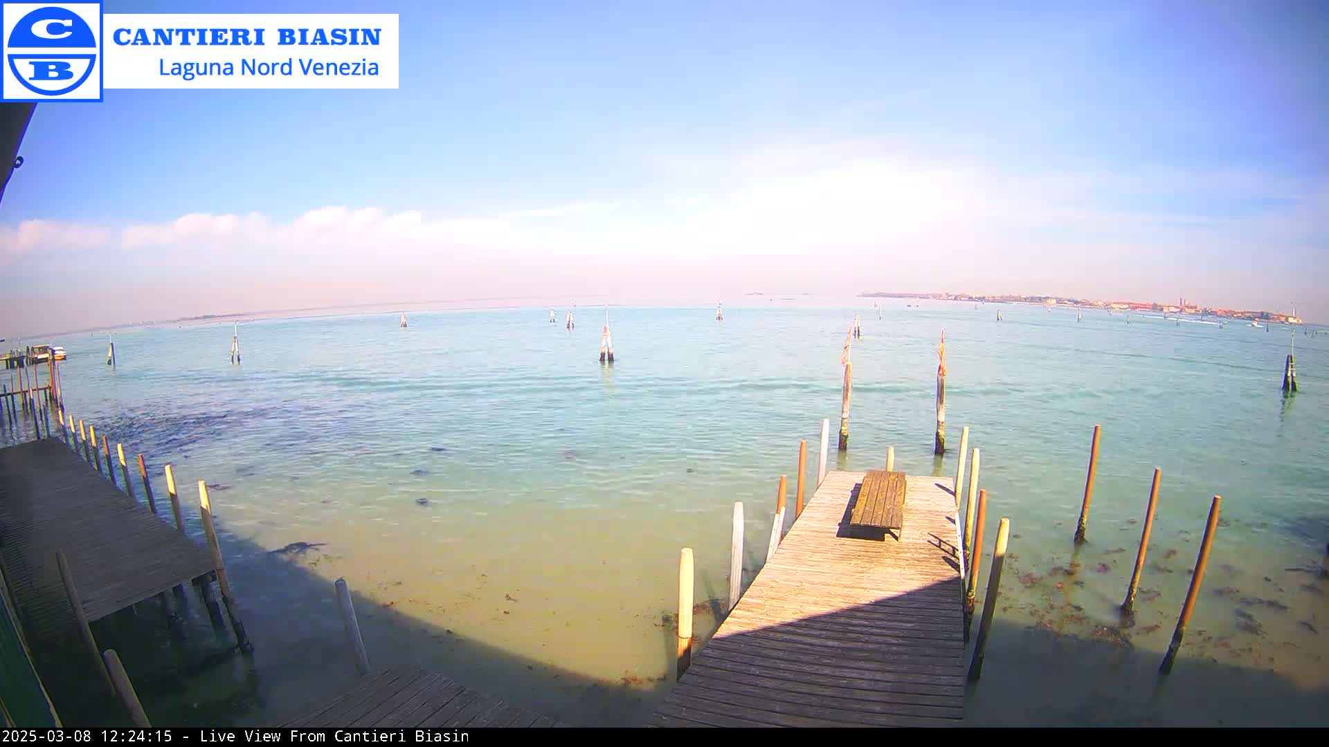 A wooden dock extends over shallow, calm, light teal water toward a distant shoreline on a sunny day with few clouds.