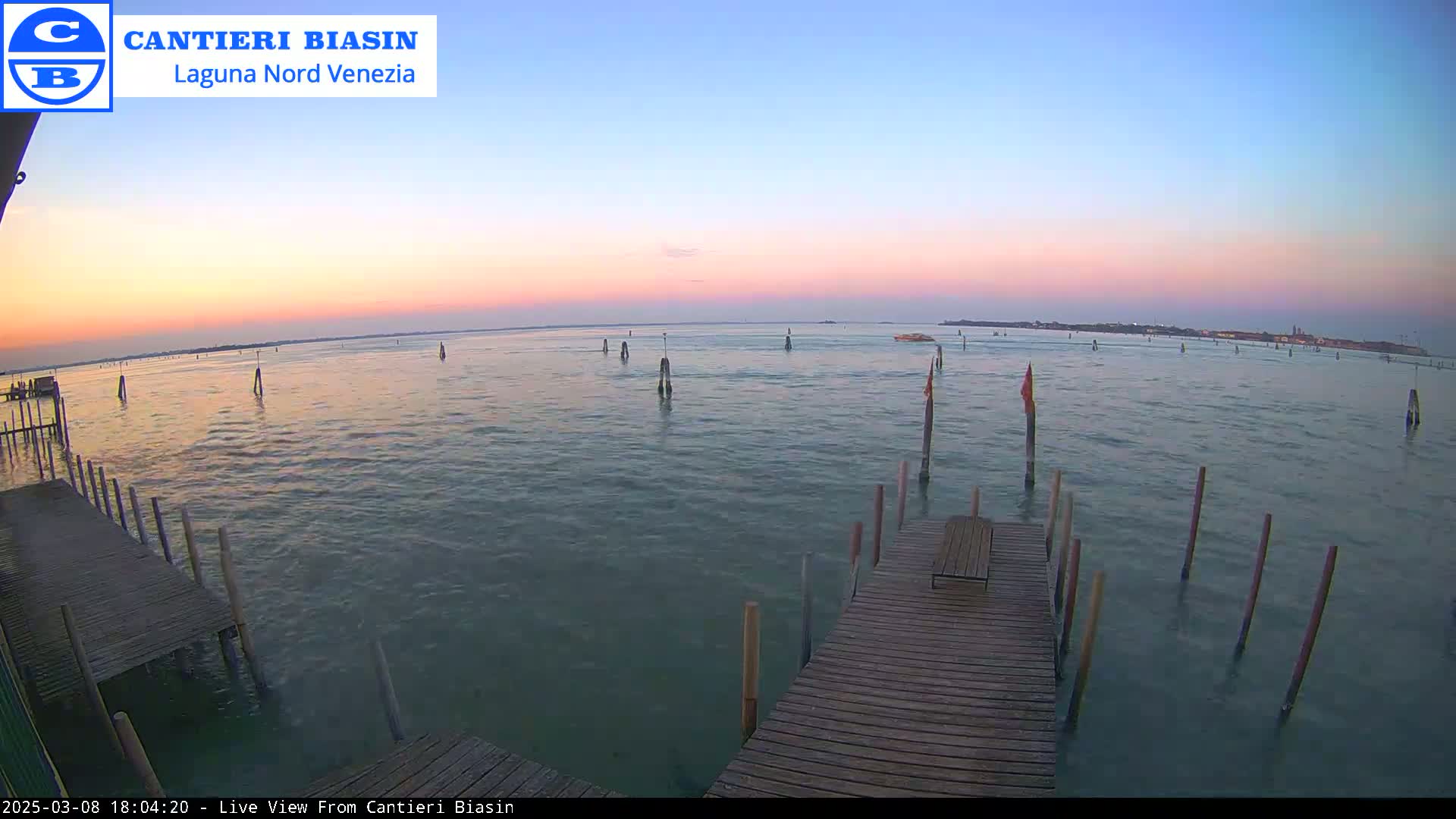 A wooden dock extends over calm water at sunset, with numerous posts visible in the water and a distant shoreline.