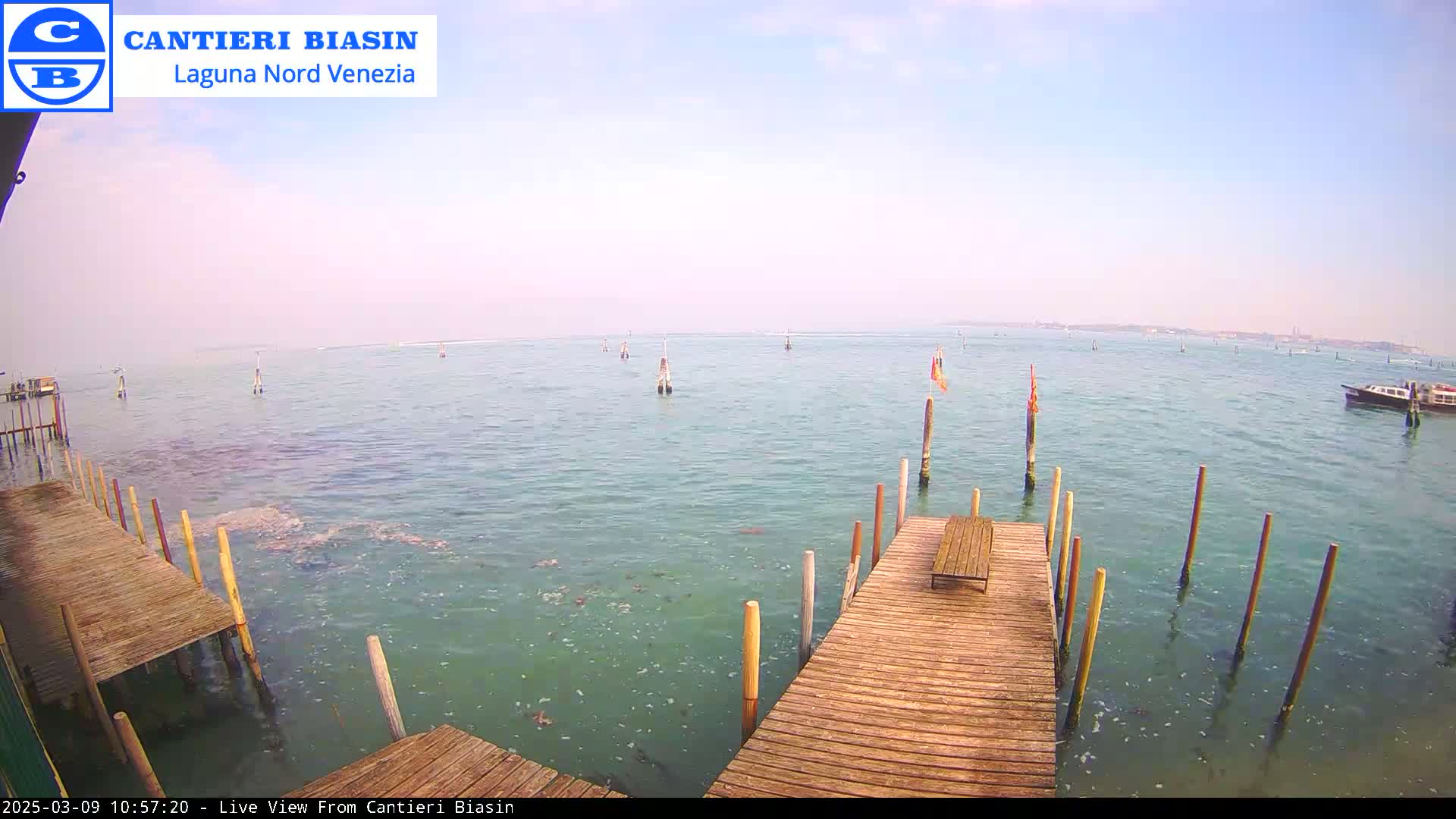 A wooden dock extends over calm, greenish-blue water, with several poles and a distant boat visible under a hazy sky.