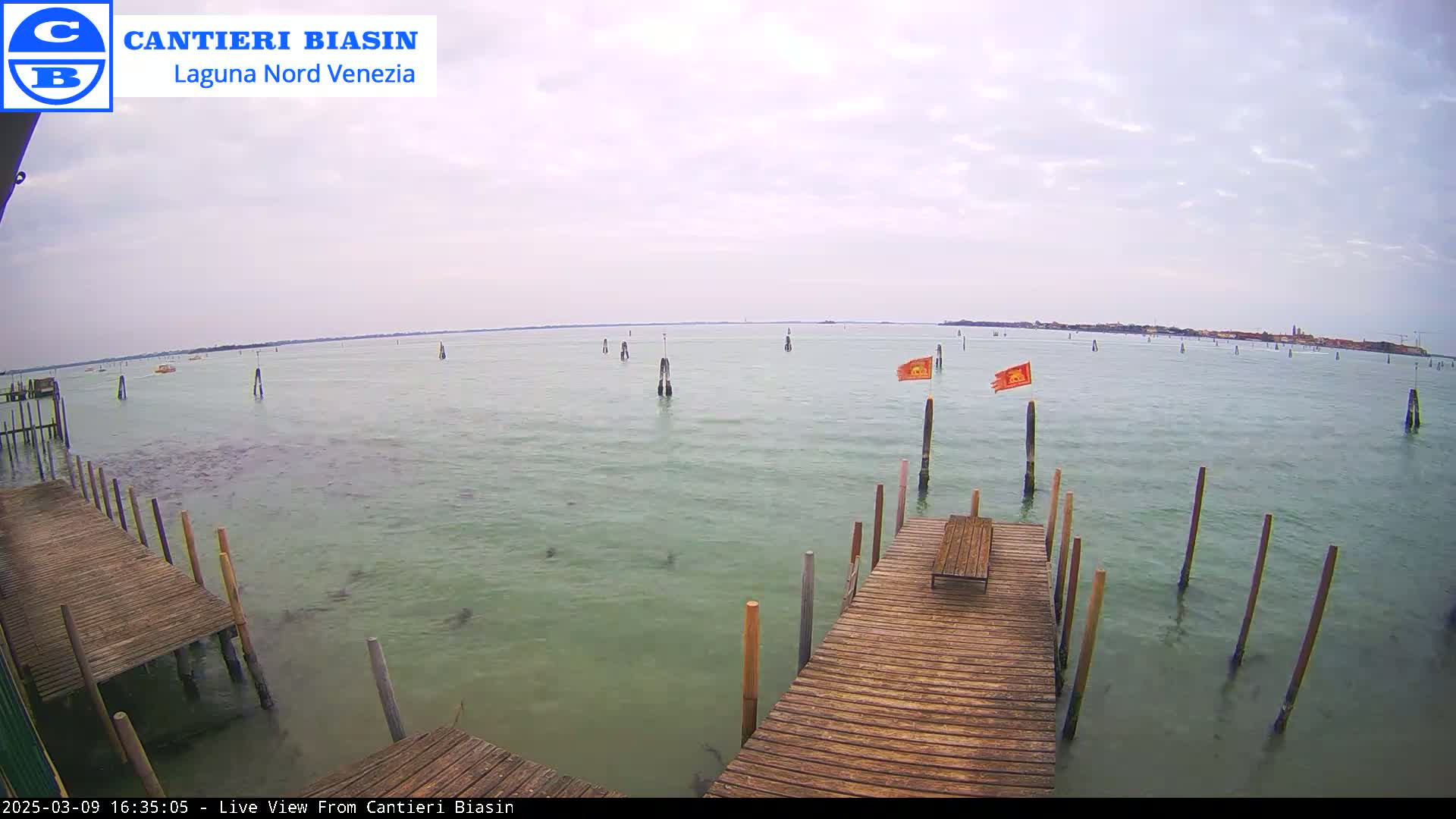 A wooden dock extends over calm, greenish-gray water under an overcast sky, with numerous wooden posts and a few orange flags visible in the distance.