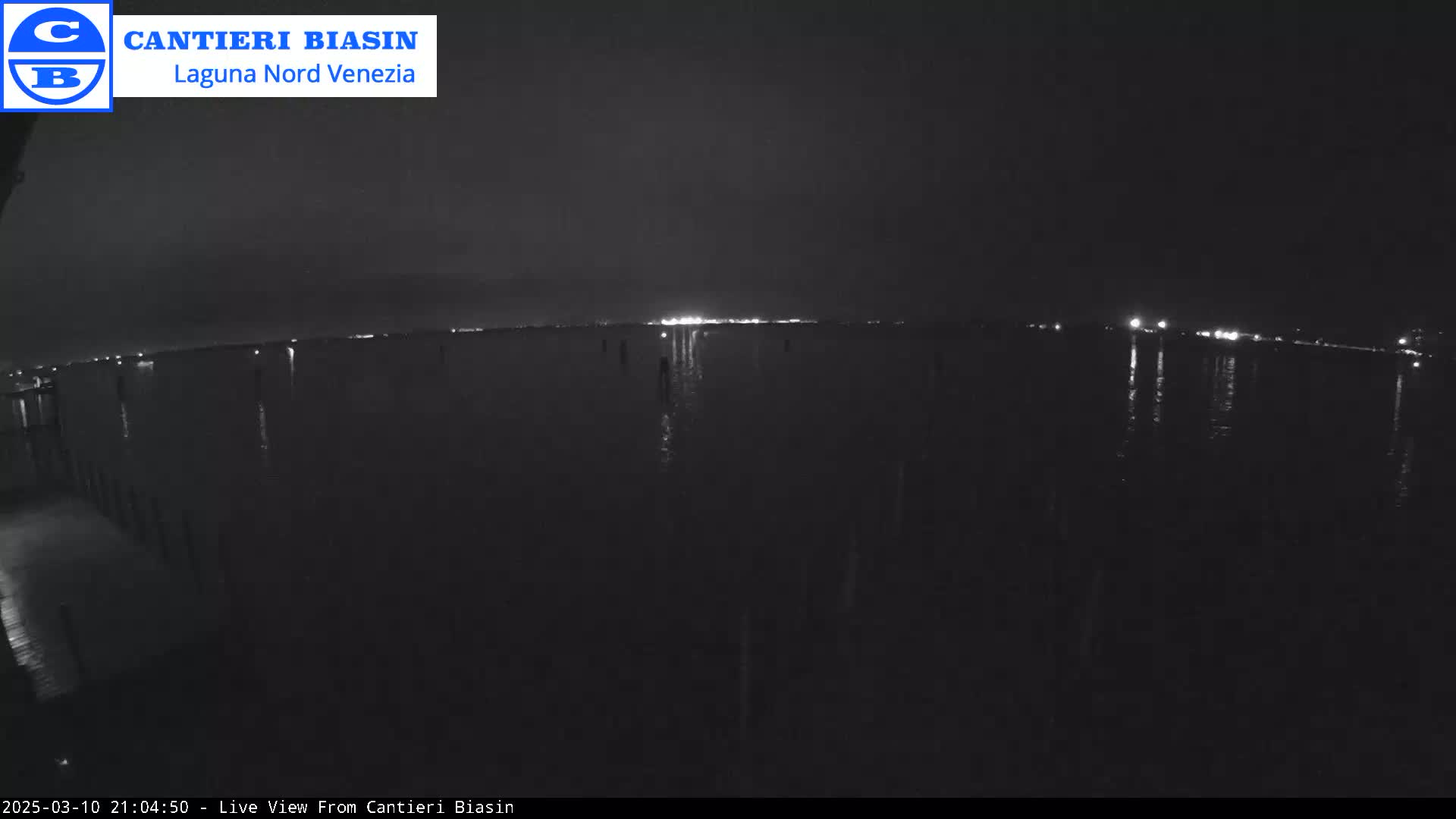 A dark nighttime view of calm water reflecting distant lights under a clear sky.