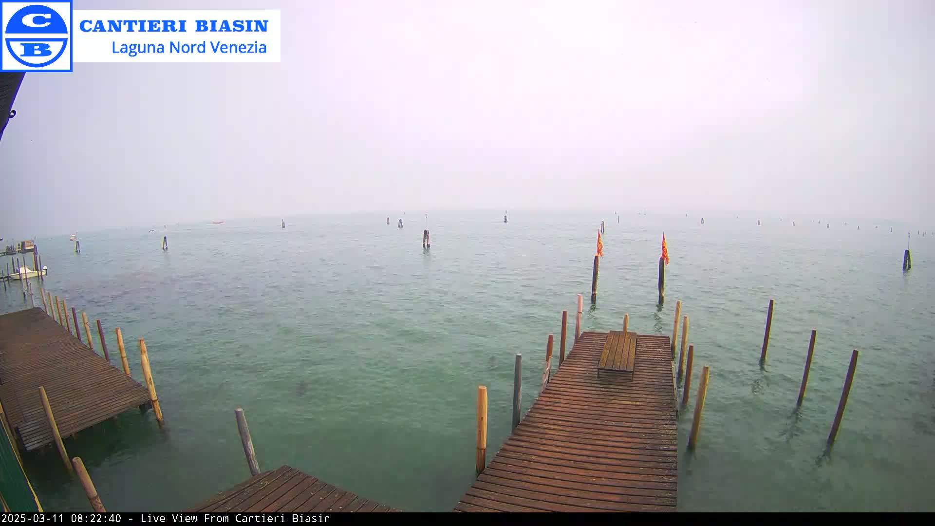 A wooden dock extends over calm, greenish water dotted with wooden posts and a few small flags, under a hazy sky.