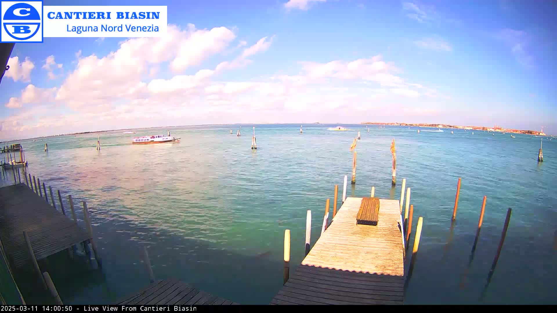 A wooden dock extends over calm, turquoise water under a mostly sunny sky with scattered clouds, a boat is visible in the distance, and land is seen on the horizon.