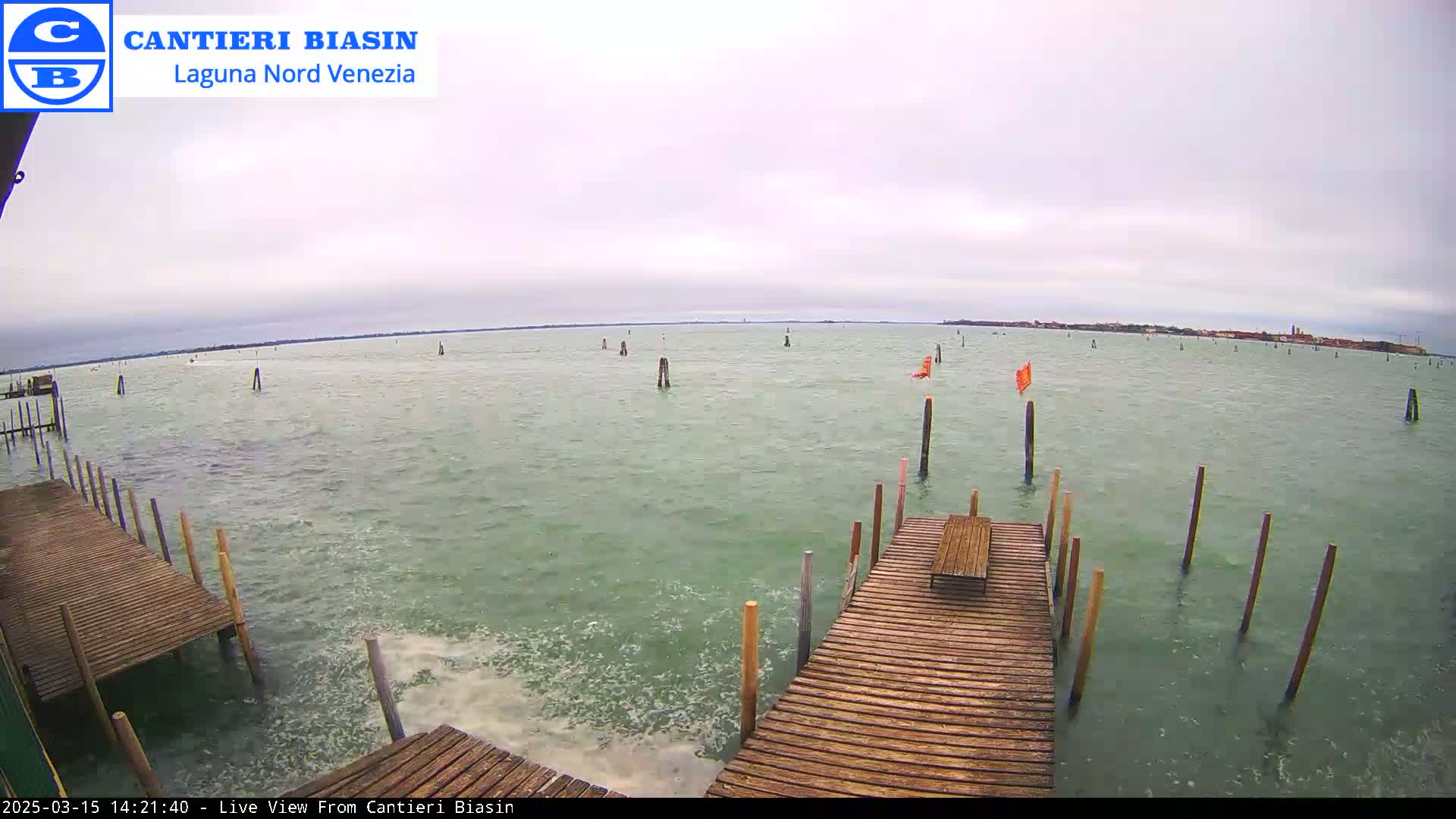 A wooden dock extends over a choppy, green body of water under an overcast sky, with numerous posts and a distant shoreline visible.