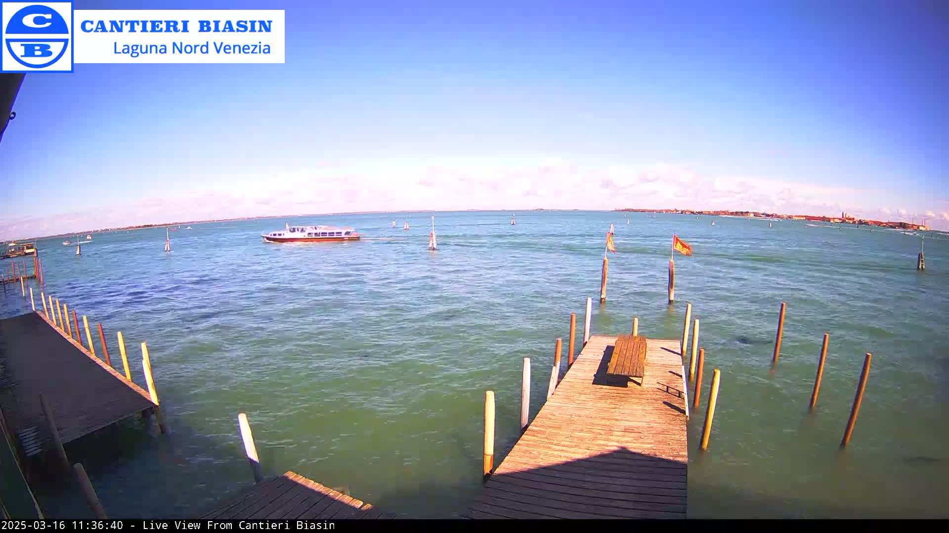 A wooden dock extends over calm, greenish water, with a small boat visible in the distance under a clear, bright sky.