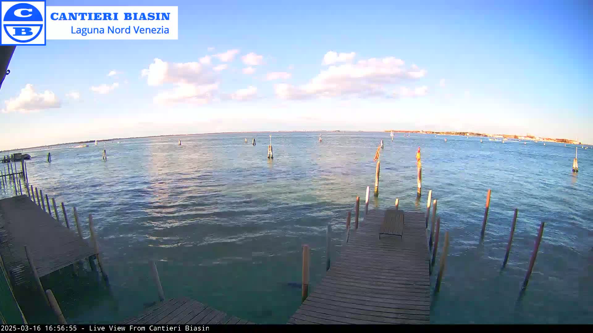 A wooden dock extends over calm, blue-green water under a mostly sunny sky with scattered clouds, toward a distant shoreline.