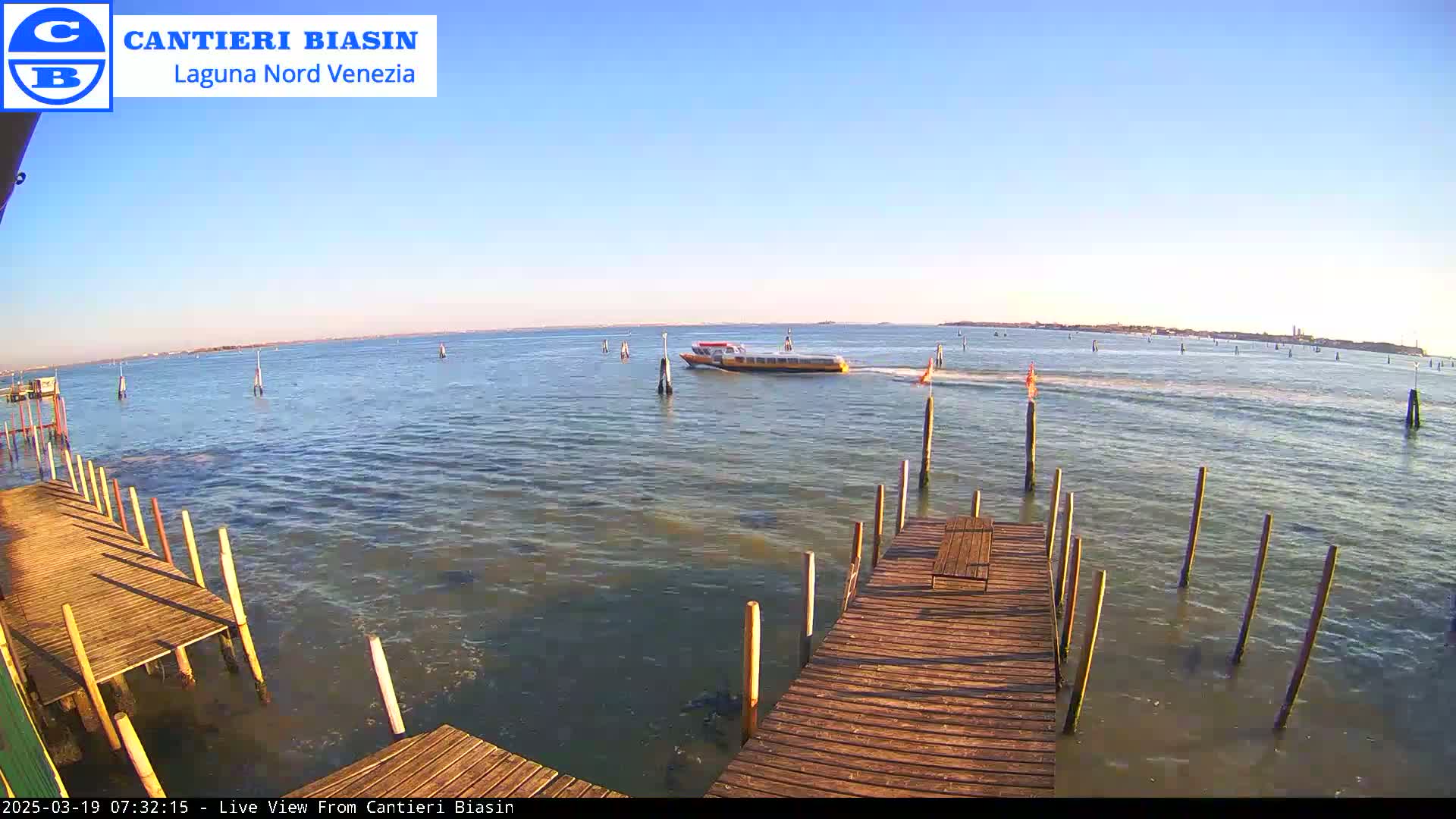 A yellow water taxi travels across calm, sunlit water toward a distant shoreline, passing wooden docks extending from the foreground.