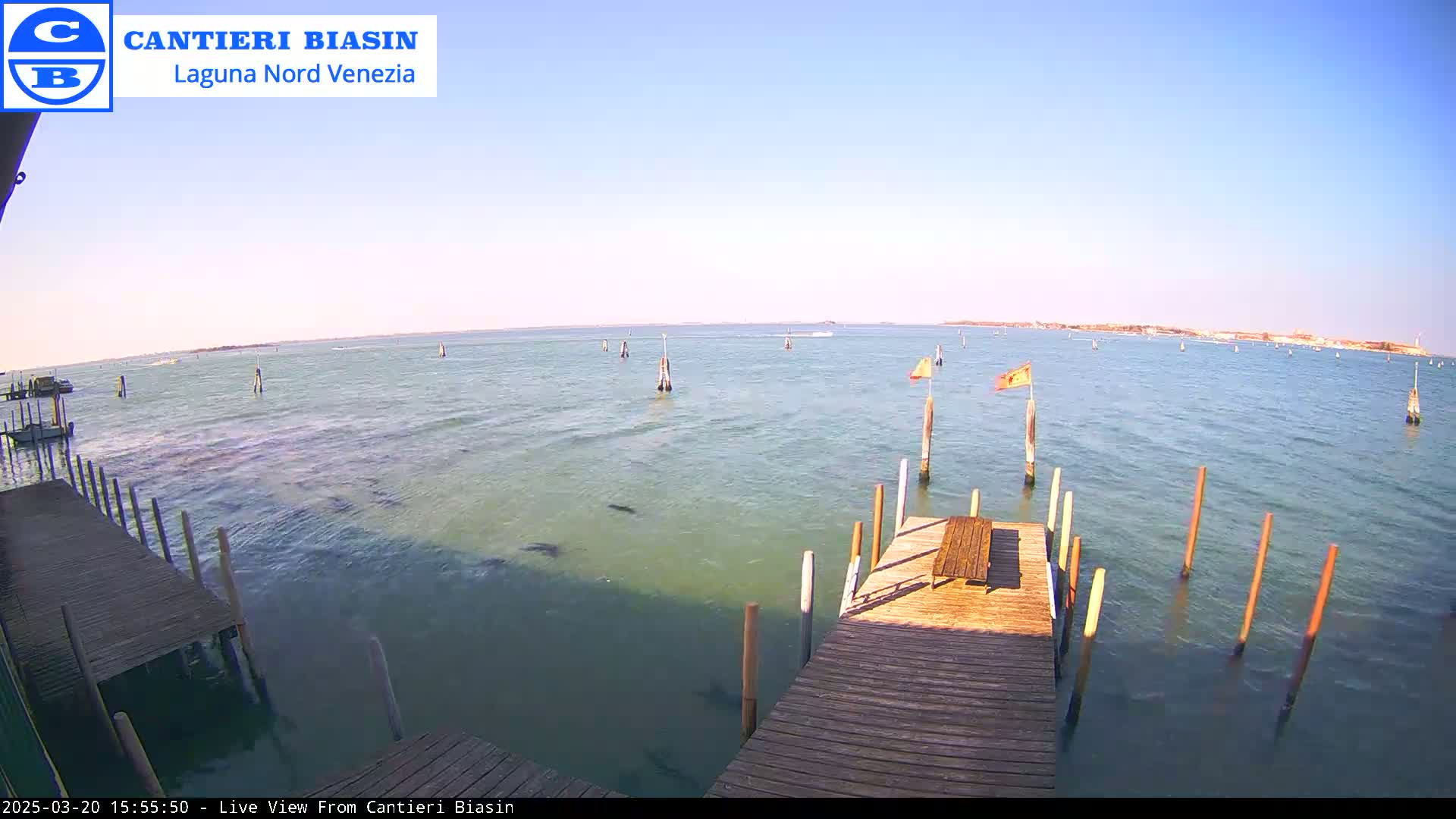 A wooden dock extends over calm, blue-green water, under a clear, bright sky;  in the distance, land is visible.