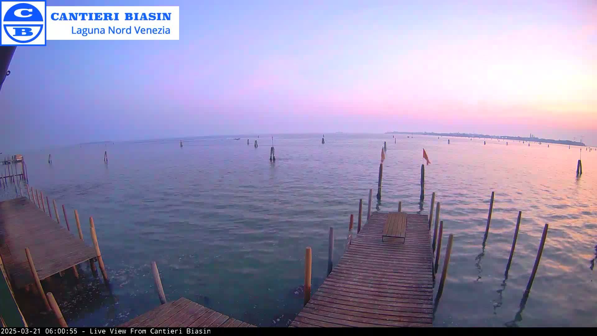 A wooden dock extends over calm water at sunset, with numerous posts and flags visible in the distance.