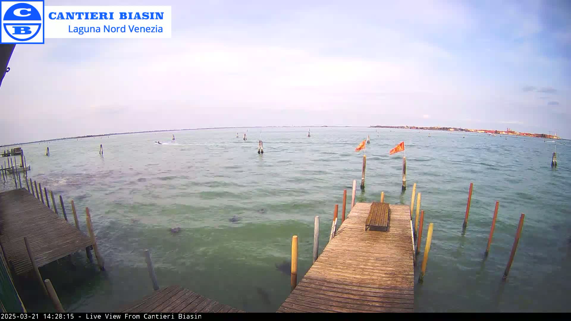 A wooden dock extends over calm, greenish-blue water with several posts and small orange flags visible, under an overcast sky.