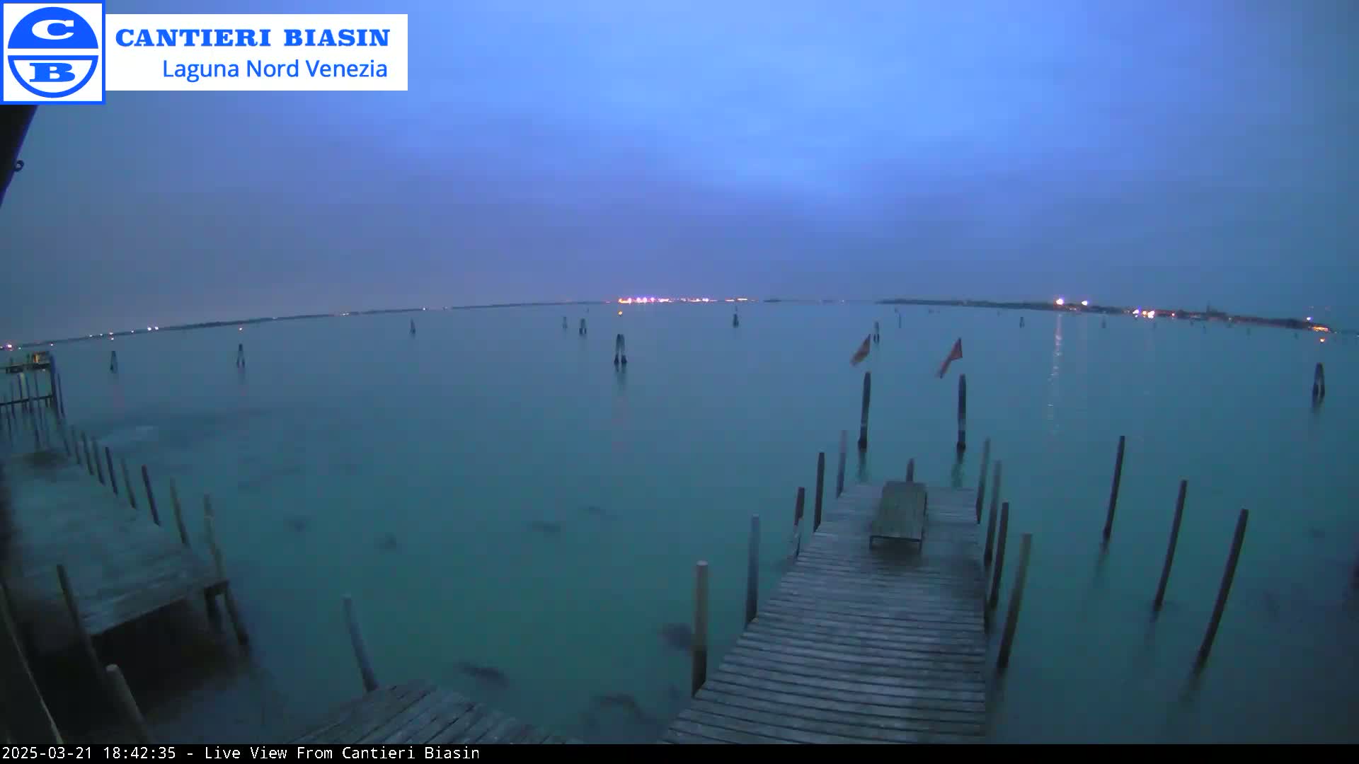 A long wooden dock extends over calm, teal water at dusk, with several posts in the water and lights visible on the distant shore.