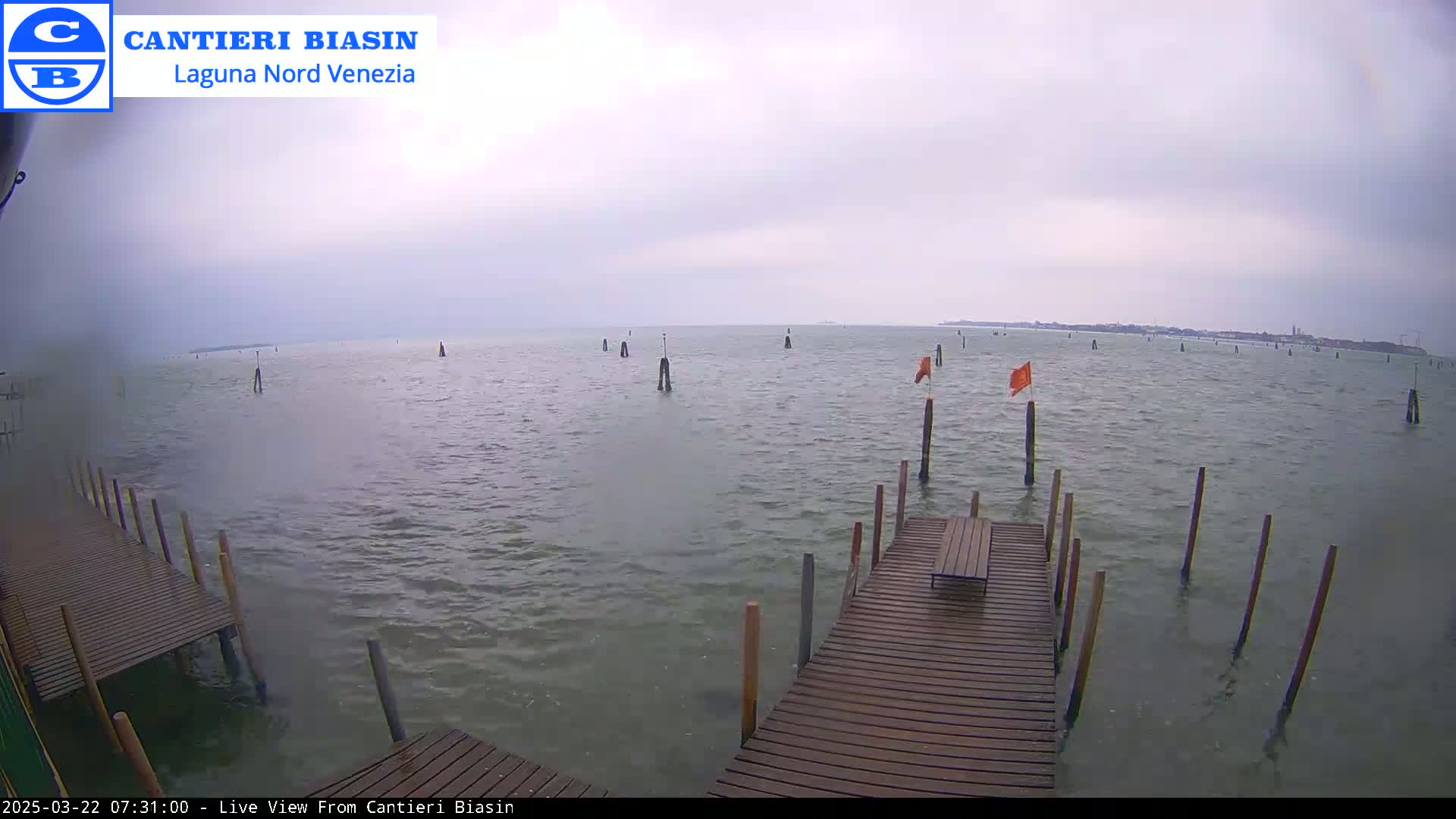 A wooden dock extends over calm, gray water under an overcast sky, with several posts and orange flags visible in the distance.