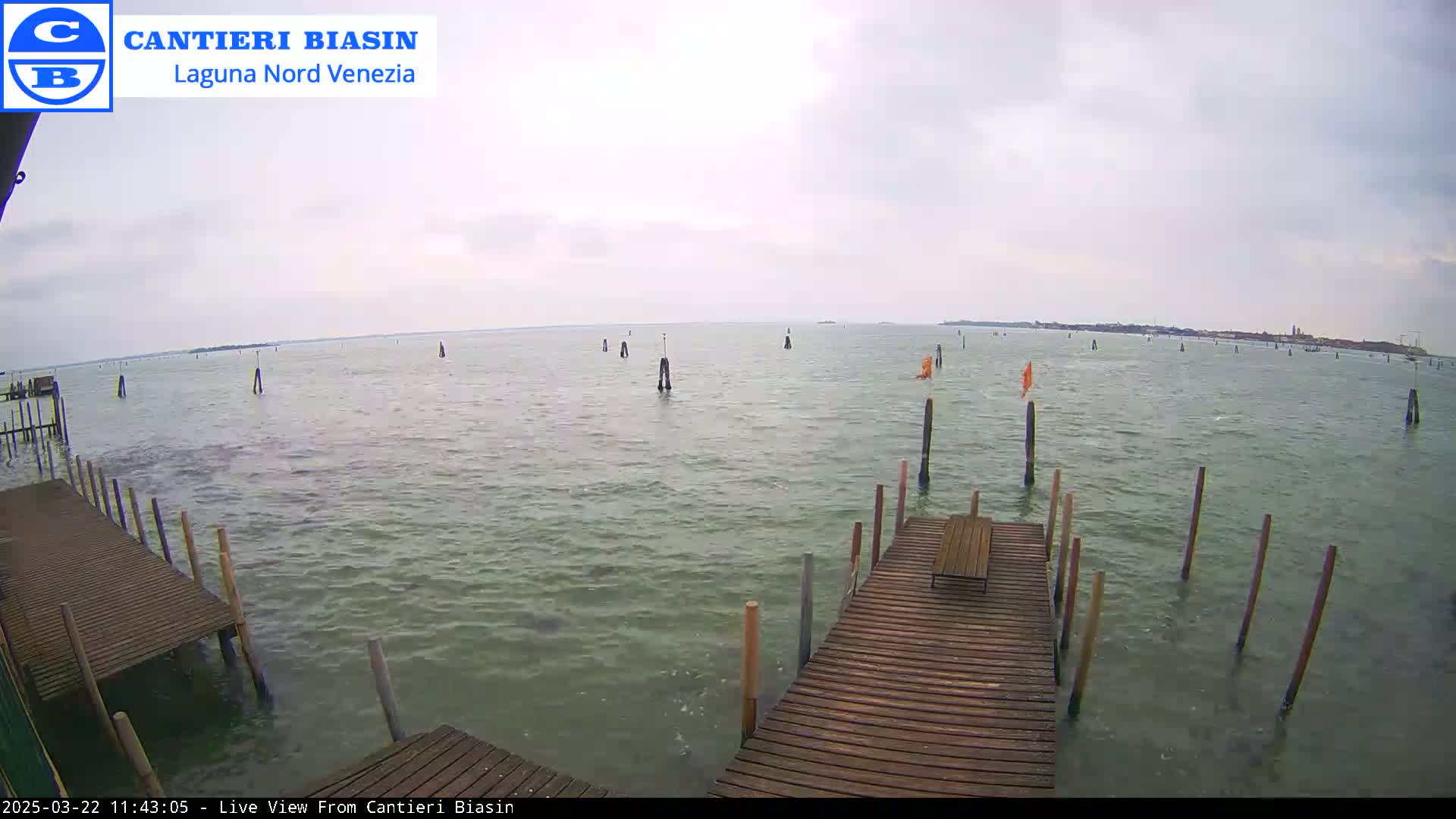 A wooden dock extends over calm, gray-green water dotted with wooden posts under an overcast sky.